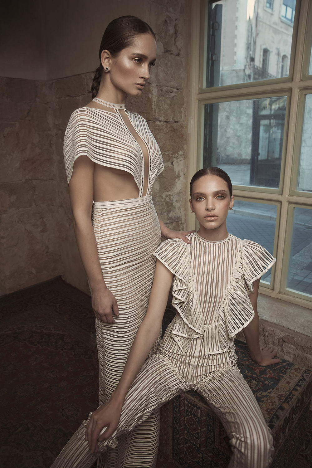 שתי נשים עם שיער חלק לאחור לובשות תלבושות אלגנטיות עם פסים בצבע שמנת — טיאנה ויארה — מקולקציית הכלות 2018, האחת בשמלה עם חיתוכים והשנייה במכנסיים עם קפלים, מצטלמות בחדר כפרי עם חלון גדול וקירות אבן.