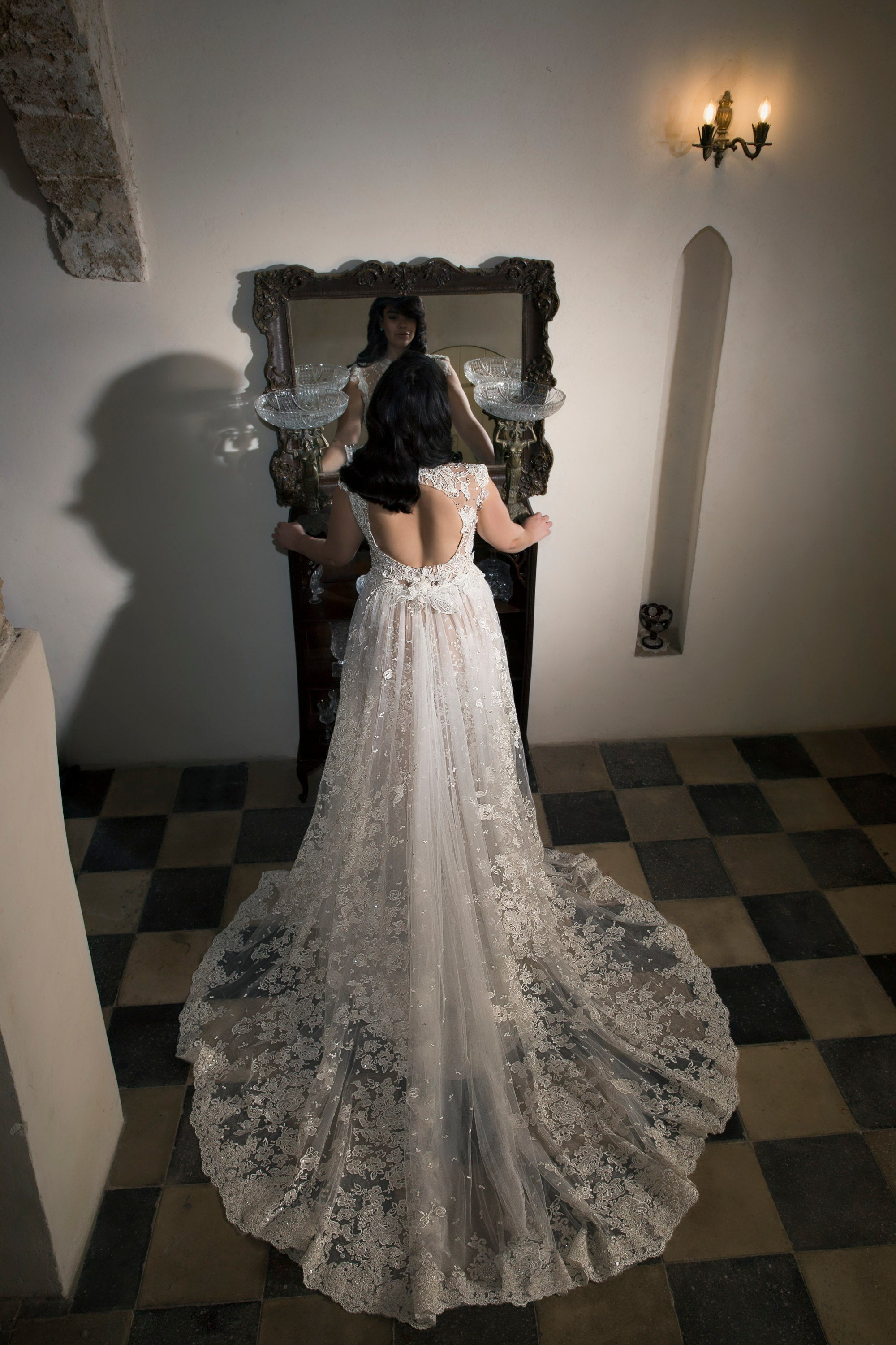 אישה לבושה בשמלת תחרה אלגנטית וחסרת גב מקולקציית Bridal Plus 2017 עומדת מול מראה על רצפה משובצת, ידיה מונחות על שידה, ותאורה רכה ופמוט קיר מאירים את הסצנה.