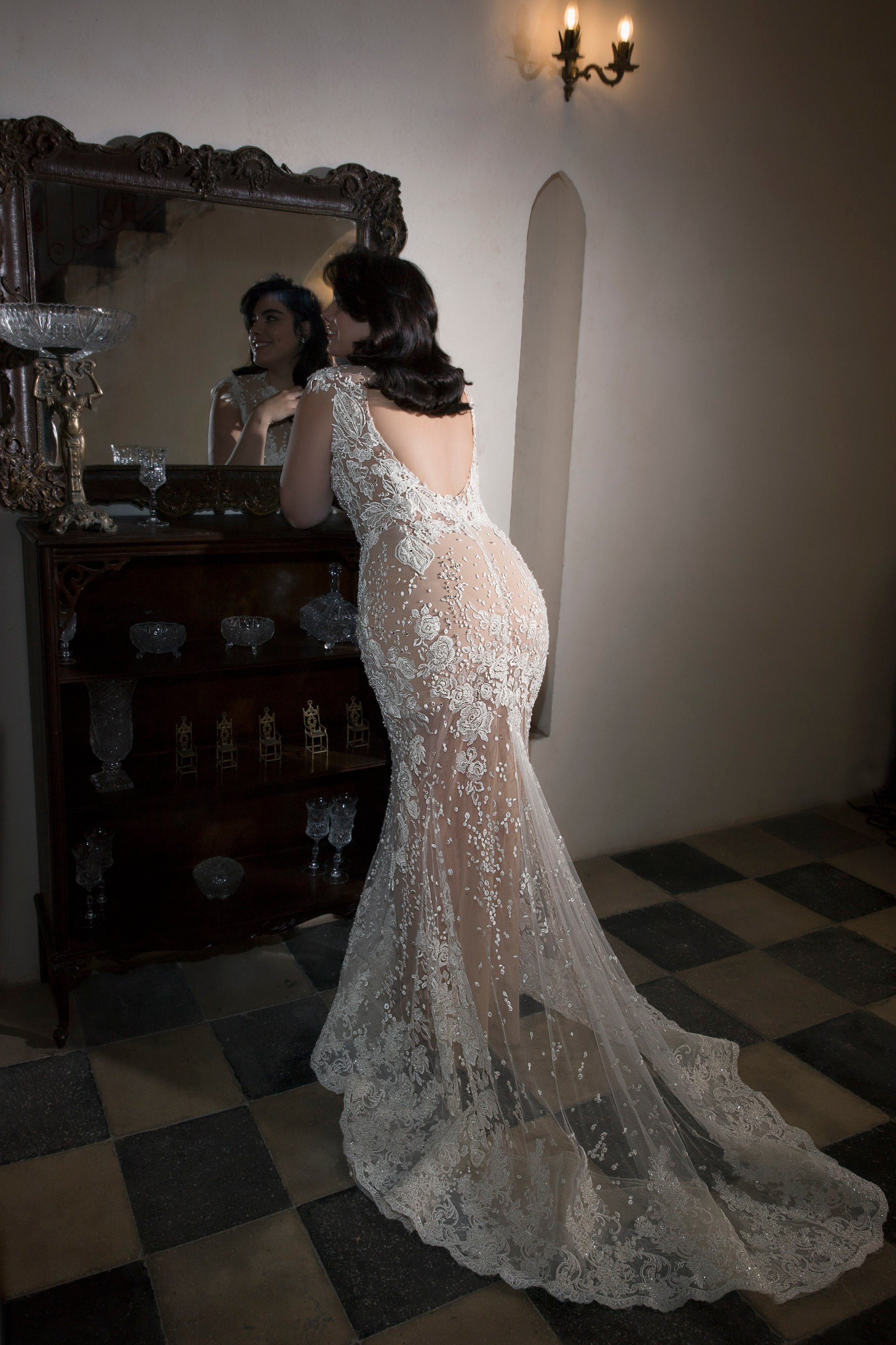 אישה בשמלת תחרה שקופה ומקושטת מקולקציית Bridal Plus 2017 - Simone by Dror Kontento עומדת ליד שידה וינטג' עם מראה, מביטה בבבואתה בחדר מואר באור עמום עם רצפת אריחים וקיר עם פמוטים.