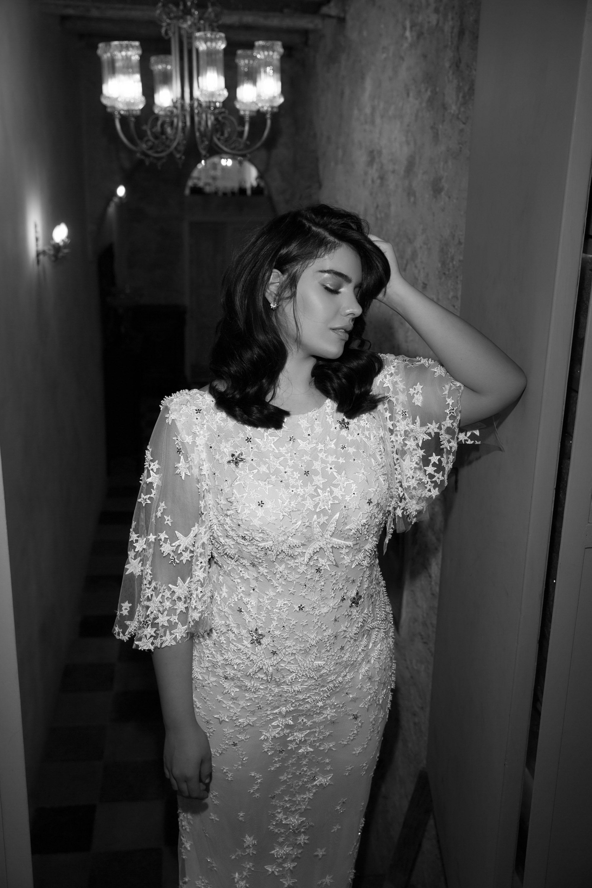 אישה בשמלת "סטאר" מקולקציית Bridal Plus 2017 עומדת במסדרון צר, עיניה עצומות, יד אחת על ראשה. תאורה רכה ונברשות וינטג' זוהרות מאחוריה בתמונה בשחור-לבן זו.