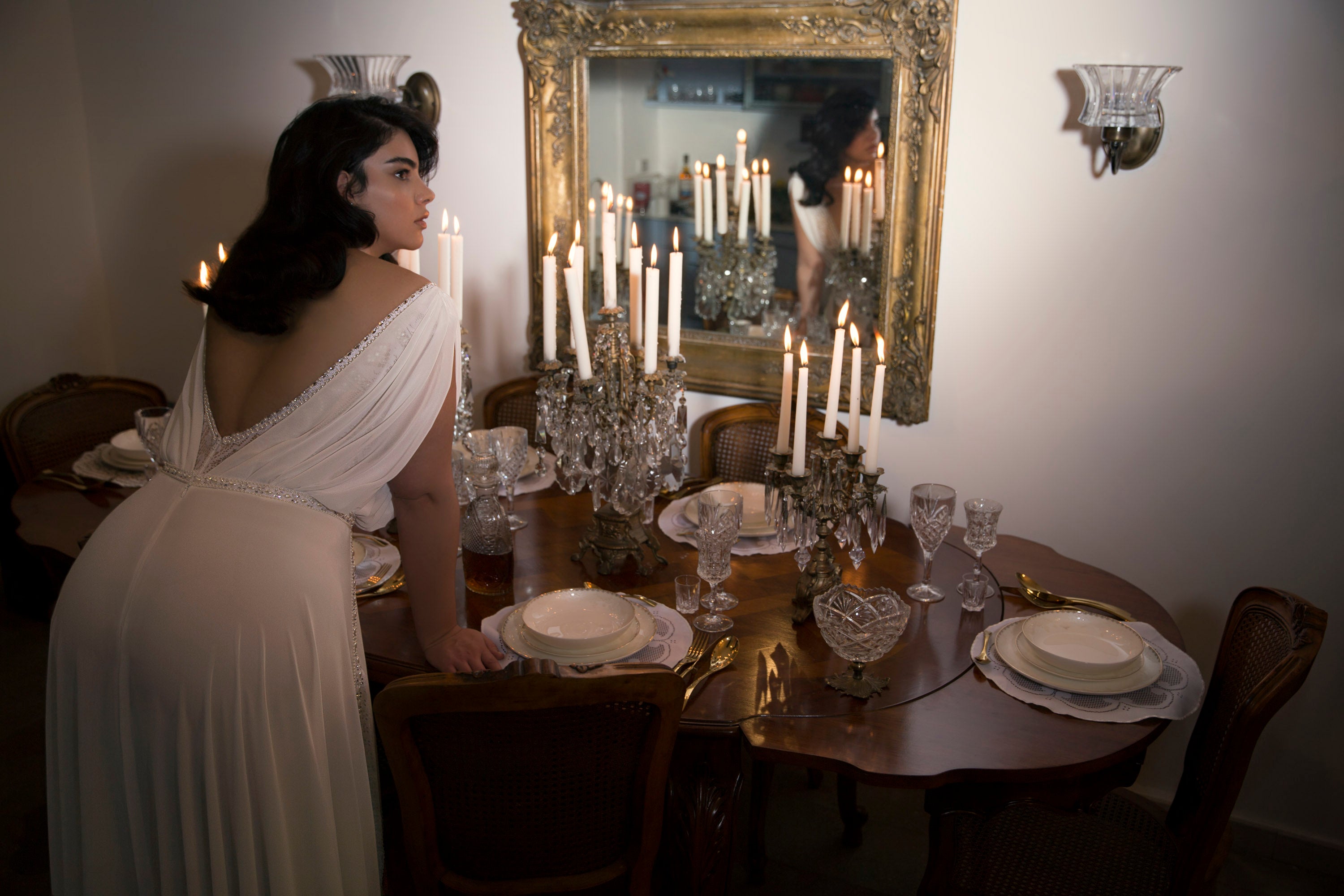 אישה בשמלה לבנה מקולקציית Bridal Plus 2017 - Saigon נשענת על שולחן אוכל ערוך עם נרות דולקים, בעוד מראה גדולה ומקושטת משקפת אותה ואת אור הנרות, ומוסיפה מגע רשמי ווינטג'י.