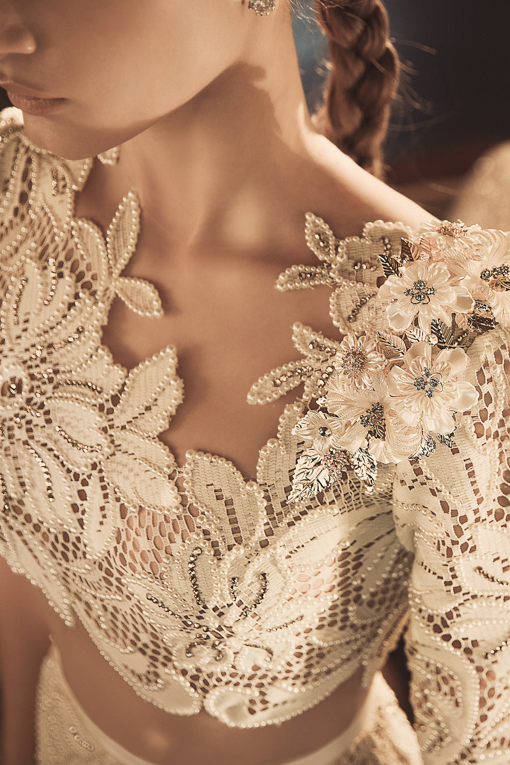 תקריב של אישה לבושה בשמלת תחרה מורכבת מקולקציית הכלות 2018 - Sunny's, עם רקמת פרחים, עבודת חרוזים ופרחים תלת-ממדיים עדינים על הכתפיים. רק חלק מפניה ופלג גופה העליון נראים.