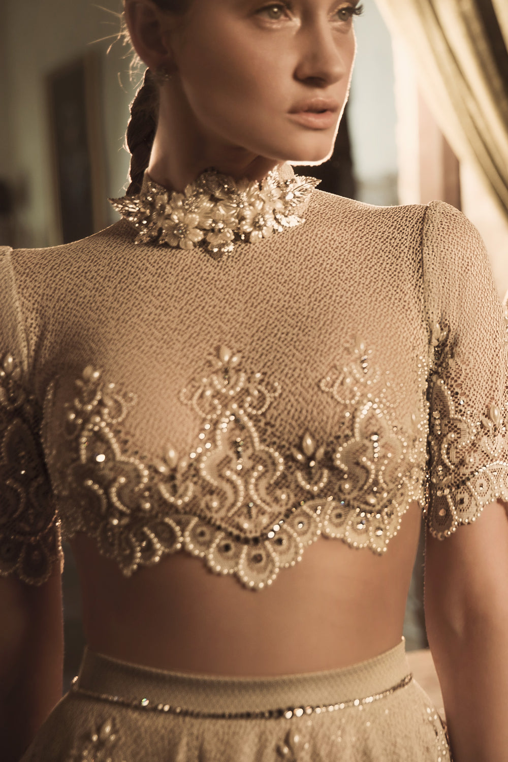 אישה עומדת באור רך וחם, לבושה בחולצת בטן קצרה מדגם "Blaire + Margo" מקולקציית הכלות 2018 של דרור קונטנטו, עם צווארון גבוה ומקושט, שרוולים שקופים ודוגמאות מעוטרות עדינות עם פרטים נוצצים.