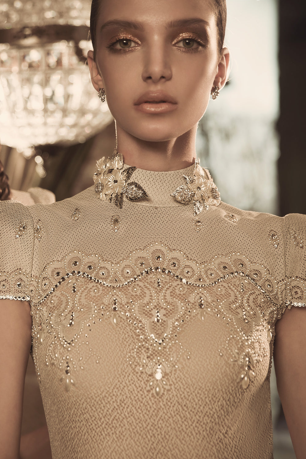 אישה לבושה בשמלת כלה מעוטרת בצבע בז' עם צווארון גבוה מקולקציית הכלות 2018 של דרור קונטנטו - Blaire + Margo, מעוטרת בחרוזים מורכבים וקישוטים נוצצים, עומדת על רקע מואר בעדינות עם איפור ותכשיטים אלגנטיים.
