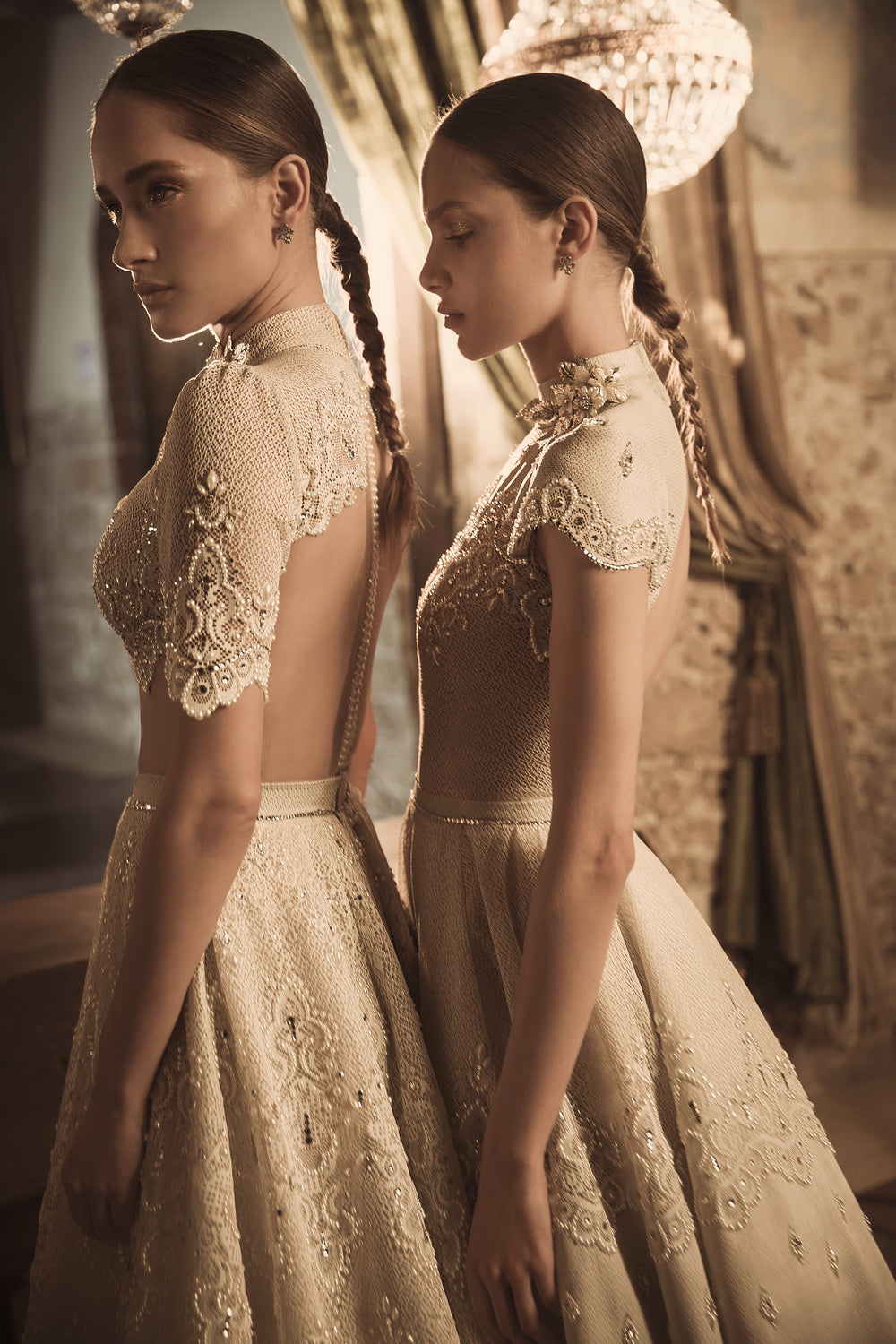 שתי נשים עם צמות ארוכות לובשות שמלות אלגנטיות מקולקציית הכלות 2018 של דרור קונטנטו - Blaire + Margo, עם תחרה וחרוזים, עומדות זו לצד זו בחדר מואר באור חם, בסגנון וינטג', עם נברשות ווילונות דראפיים.