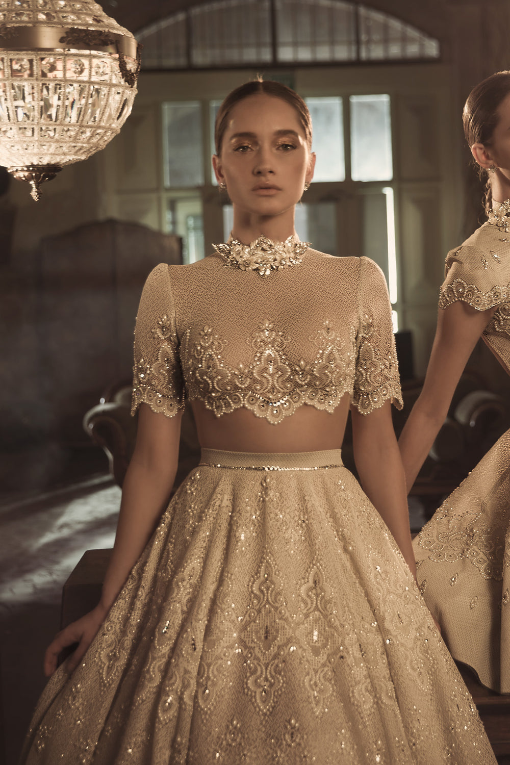 אישה עומדת בחדר וינטג' אלגנטי לבושה בשמלת Blaire + Margo מקולקציית הכלות 2018, עם רקמת חרוזים מורכבת, צווארון גבוה משובץ אבני חן ושרוולים קצרים. שיערה אסוף לאחור מתחת לנברשת נוצצת.