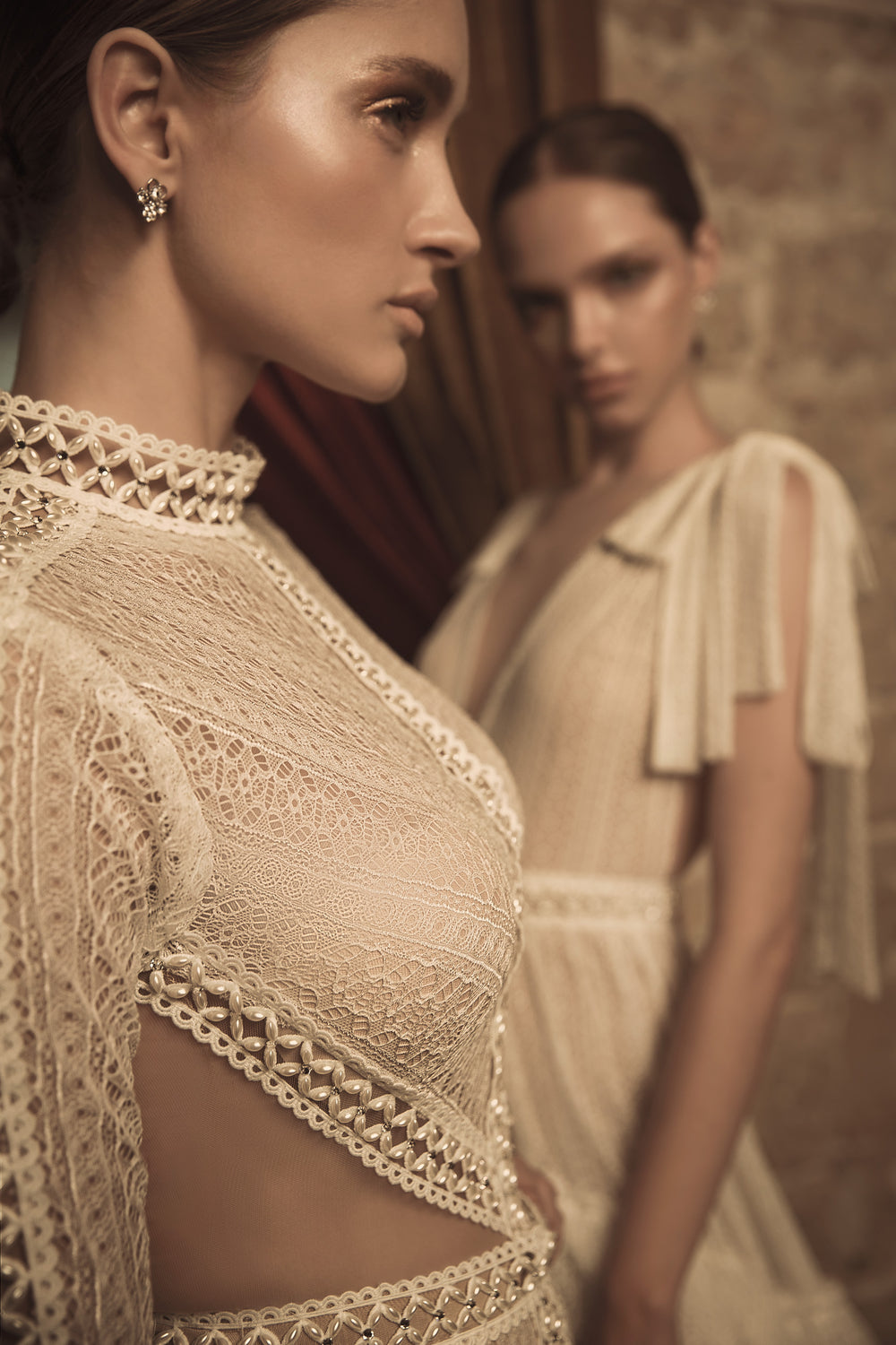 שתי נשים לובשות שמלות תחרה בצבע שמנת מקולקציית הכלות 2018 של דרור קונטנטו - Nia + Erin. האישה הקדמית ממוקדת בחדות, מה שמדגיש את פרטי הבד, בעוד שהשנייה נראית מטושטשת ברקע.