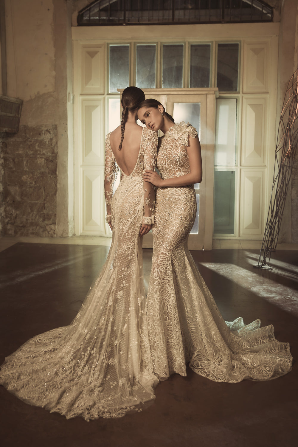 שתי נשים בשמלות אלגנטיות וארוכות מהקולקציית הכלות 2018 - Marie + Angelica עומדות זו לצד זו בחדר מואר באור רך עם דלתות לבנות גדולות וקירות כפריים, האחת מניחה את ראשה בעדינות על כתפה של השנייה.