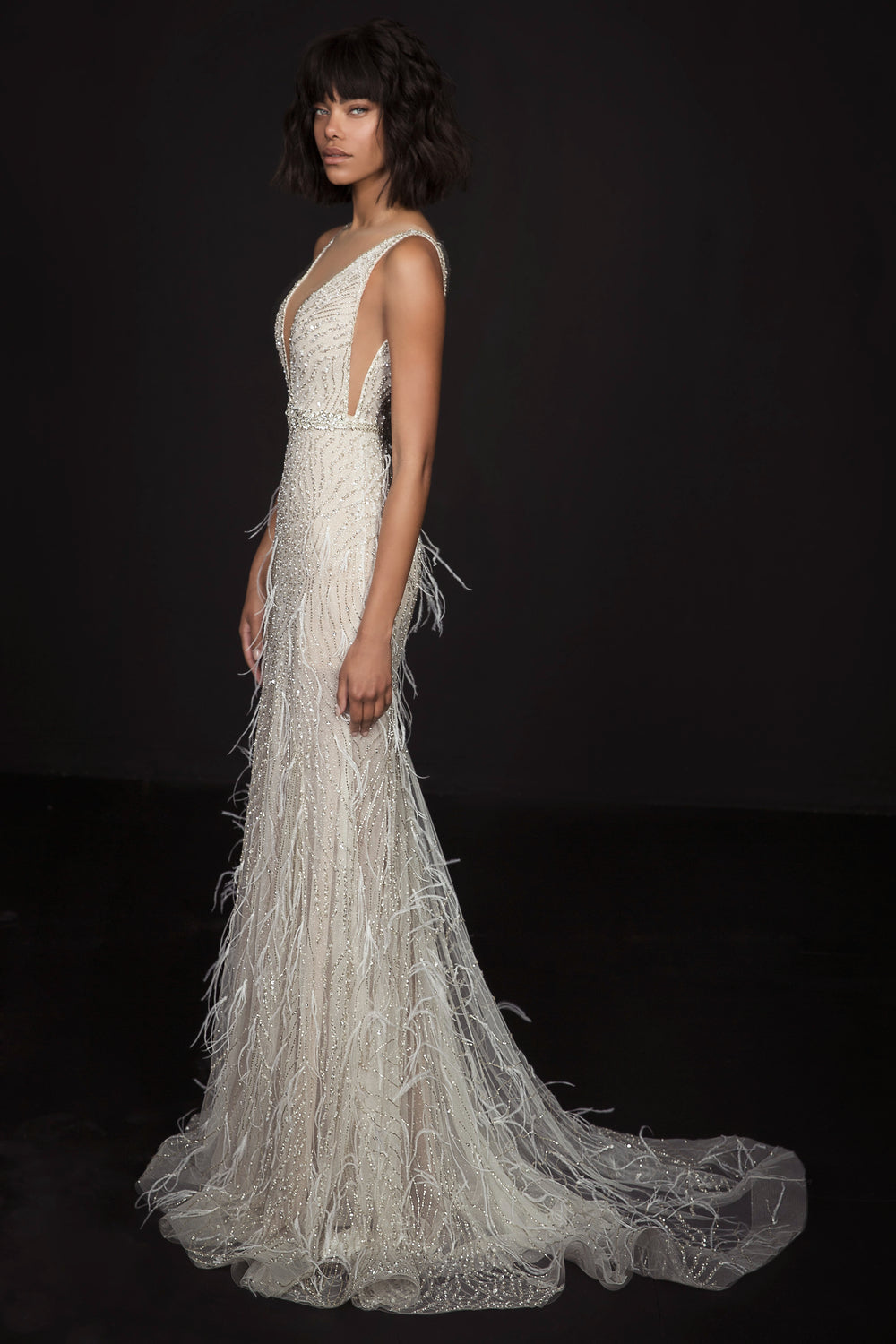 אישה עם שיער קצר וכהה מצטלמת בשמלת Romina מקולקציית הכלות 2017 של דרור קונטנטו — שמלה בצבע שנהב, ללא שרוולים, באורך הרצפה, עם עיטורי חרוזים, פרטים דמויי נוצות, מחשוף V עמוק ושובל ארוך, על רקע כהה.