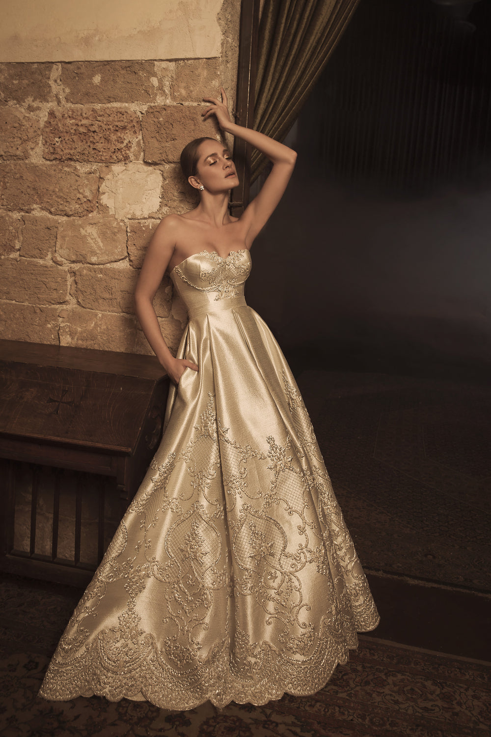 אישה בשמלת "אליס" בצבע שמפניה ללא כתפיות מקולקציית הכלות 2018 של דרור קונטנטו, מצטלמת באלגנטיות על רקע קיר אבן כפרי, עם תאורה חמה המדגישה את הרקמה המורכבת ואת פרטי שמלות הכלה.