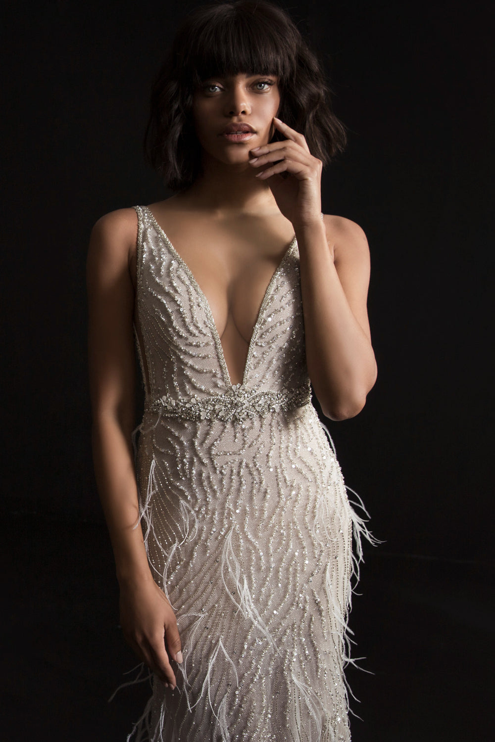 אישה עם שיער כהה באורך הכתפיים לובשת את שמלת "רומינה" מקולקציית הכלות 2017 של דרור קונטנטו, שמלה ללא שרוולים עם מחשוף V עמוק, משובצת בחרוזים עם דוגמאות מנצנצות ופרטים דמויי נוצות, על רקע כהה.