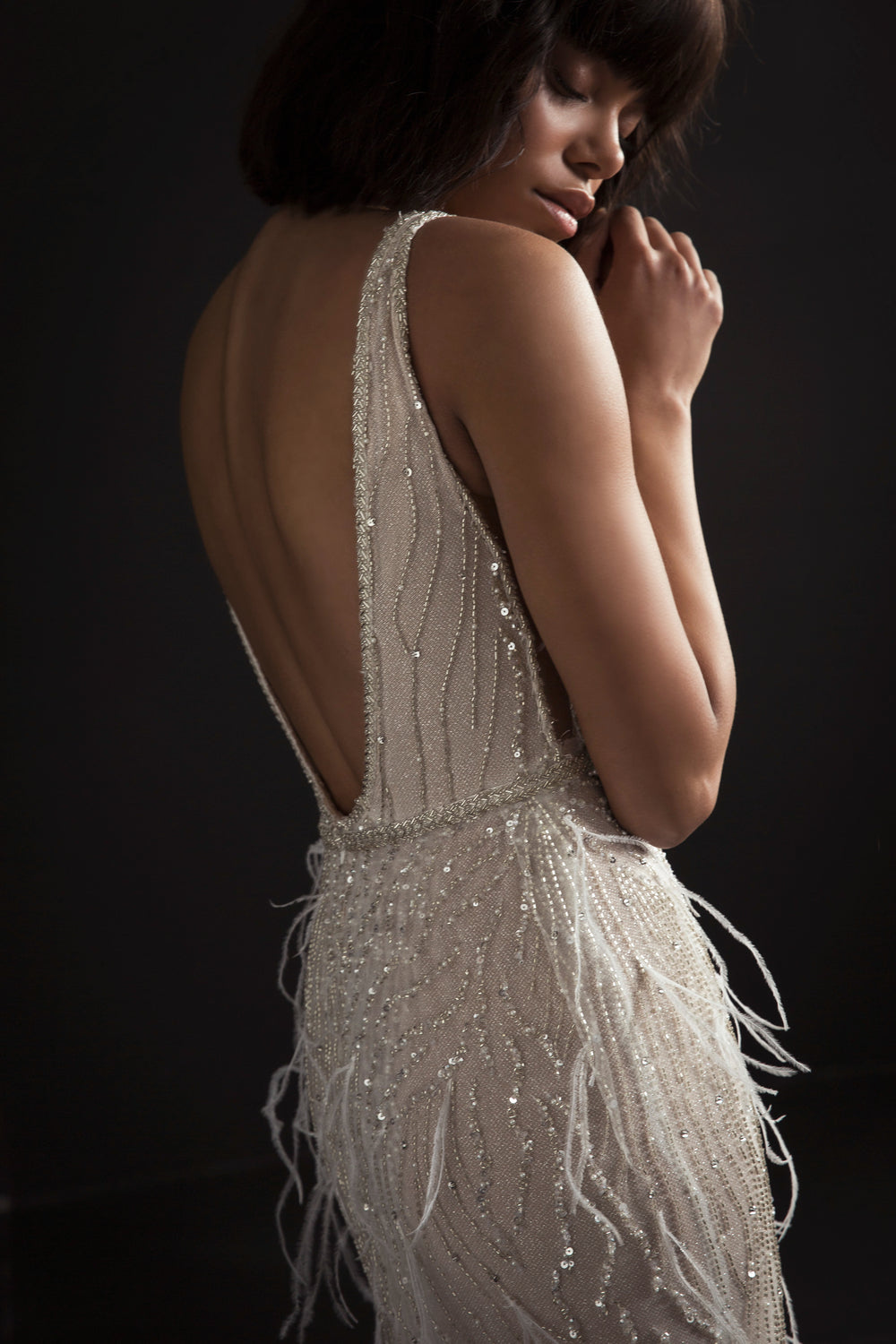 אישה בשמלת "קולקציית כלות 2017 - רומינה" של דרור קונטנטו, ללא שרוולים, נוצצת, עם עיטורי חרוזים מורכבים ופרטי נוצות, עומדת על רקע כהה, מביטה מטה, זרוע אחת מורמת בעדינות ליד פניה.