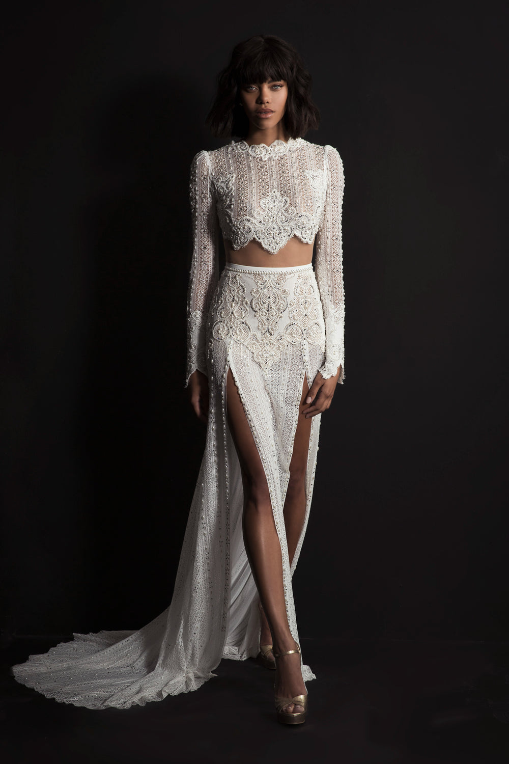 אישה מצטלמת על רקע כהה בשמלת Leyla מקולקציית הכלות 2017 של דרור קונטנטו — שמלה לבנה מעוטרת עם שרוולים ארוכים, חלק עליון קצר מתחרה, חתכים גבוהים בצדדים, שובל ועקבים מתכתיים.