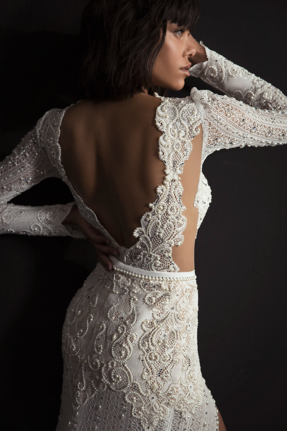 אישה לבושה בשמלת ליילה לבנה, מעוטרת בחרוזים מורכבים, עם גב פתוח ושולי תחרה מסולסלים מקולקציית הכלות 2017, עומדת על רקע כהה ומביטה לצד.