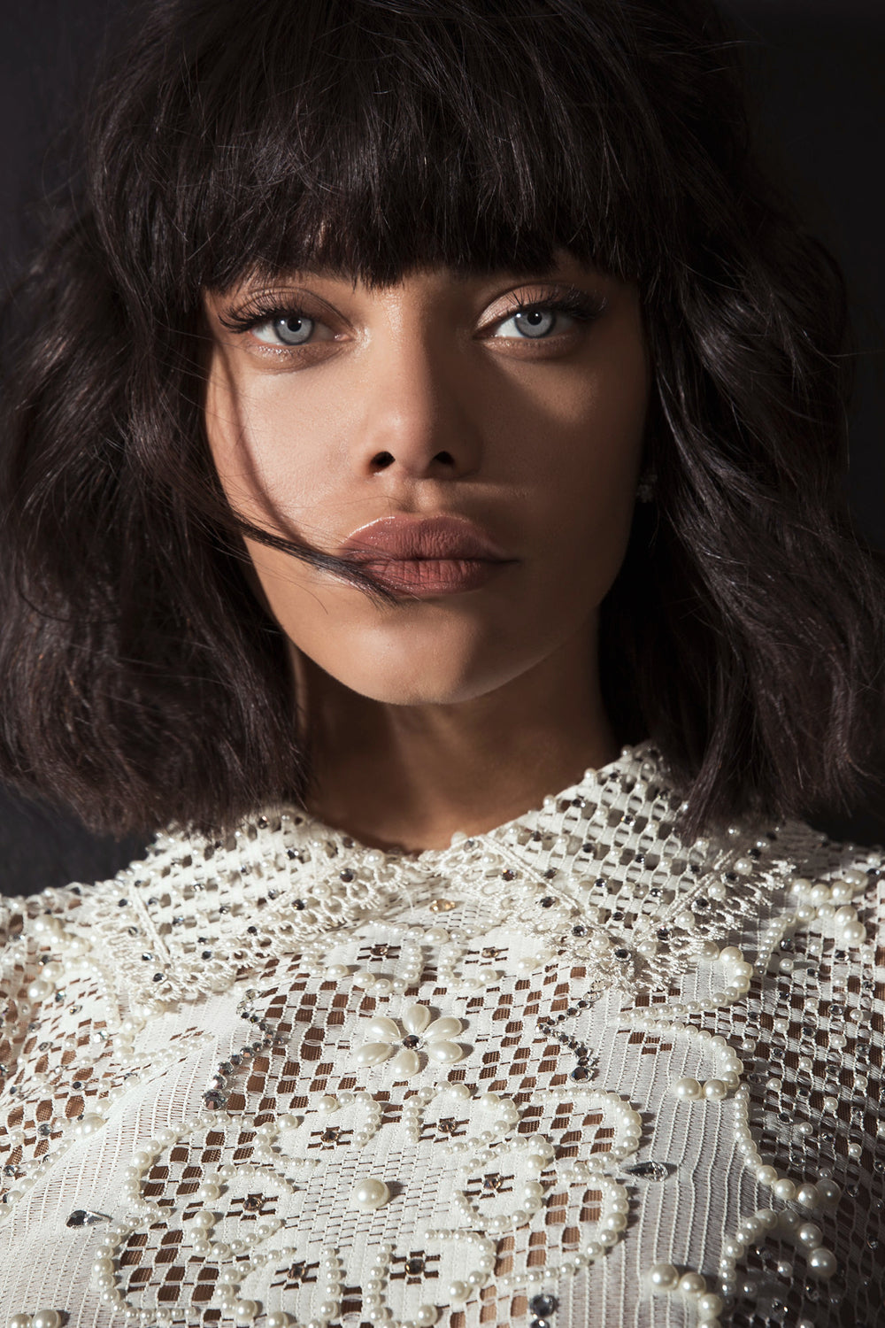 אישה עם שיער קצר וכהה ופוני לובשת את הדגם "Treasure" מקולקציית הכלות 2017 של דרור קונטנטו — חולצת תחרה לבנה עם פנינים ודוגמאות פרחוניות — ומביטה ישירות למצלמה על רקע כהה.