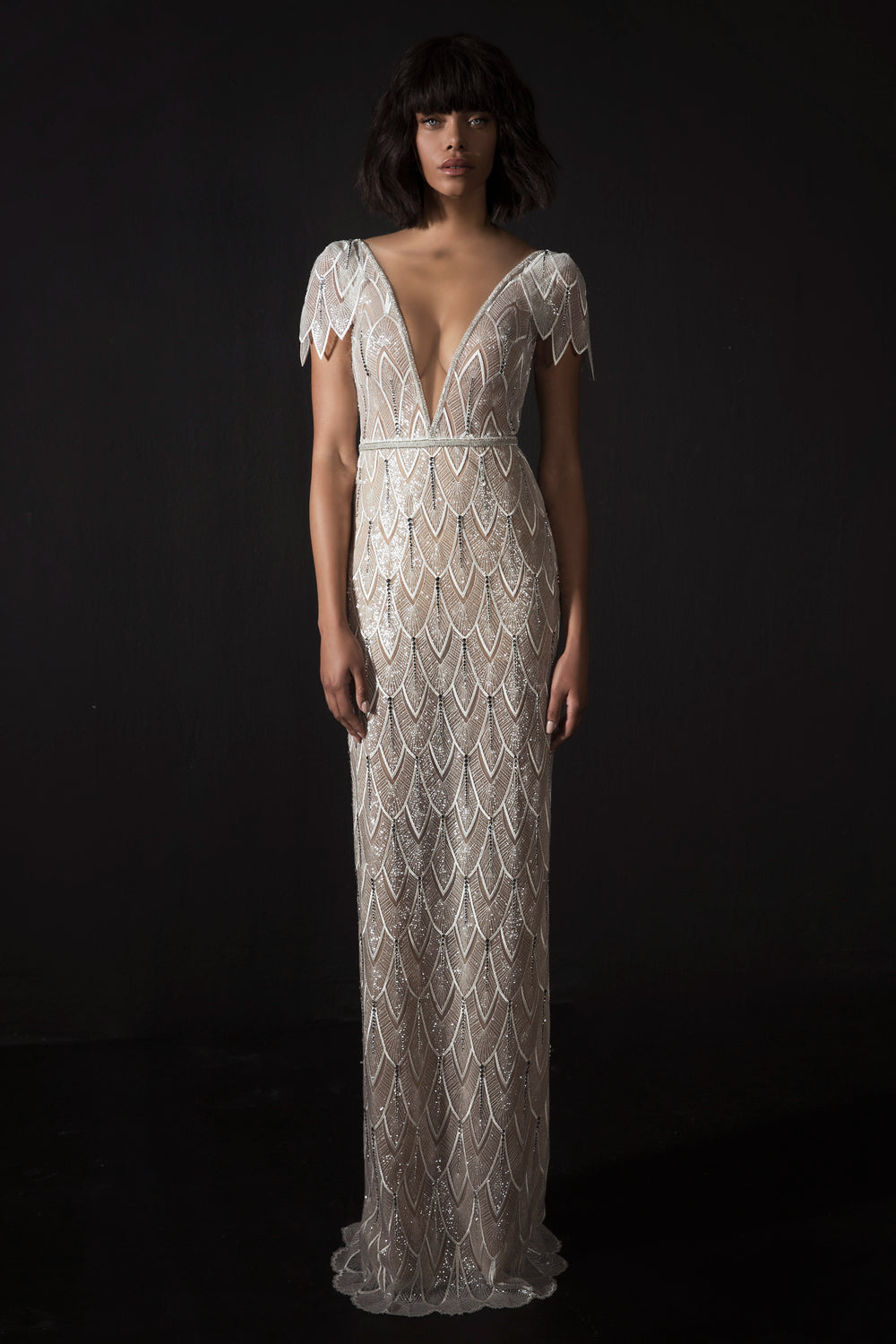 אישה עומדת על רקע כהה, לבושה בשמלת "קימבר" ארוכה ומחמיאה מקולקציית הכלות 2017 של דרור קונטנטו, עם מחשוף V עמוק, שרוולים קצרים ודוגמת סקלופס נוצצת בהשראת ארט דקו.