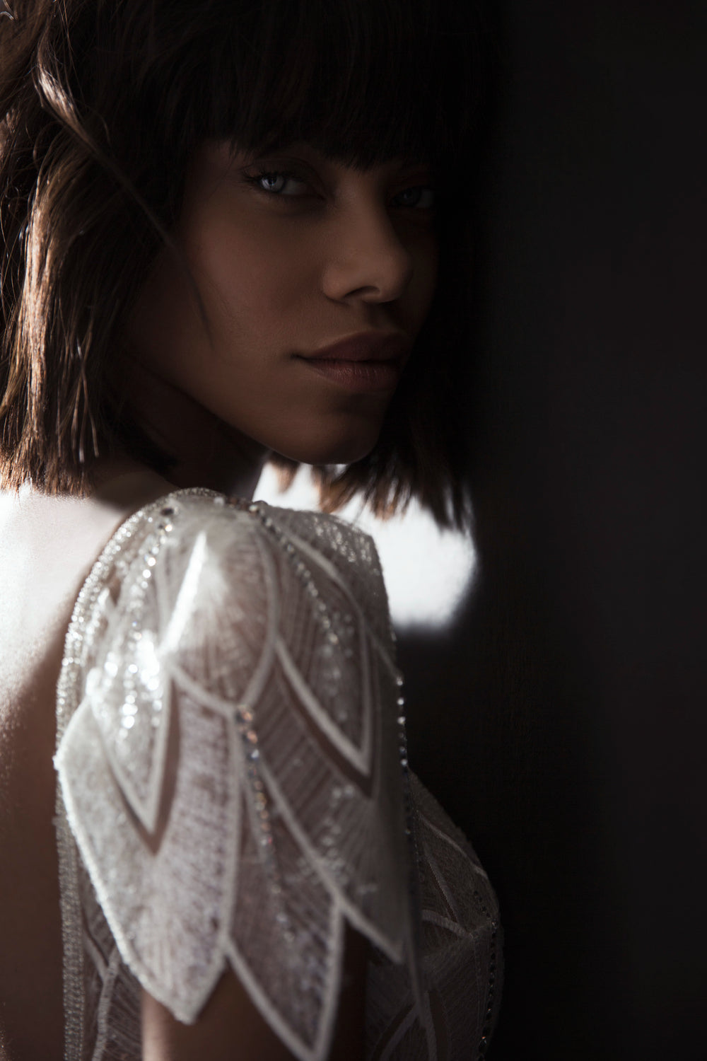 אישה עם שיער קצר כהה ופוני עומדת בצללים, מביטה במצלמה באור רך. היא לובשת את שמלת הכלה Kimber מקולקציית הכלות 2017, עם דוגמאות גיאומטריות של חרוזים המנצנצים על שמלות כלה.