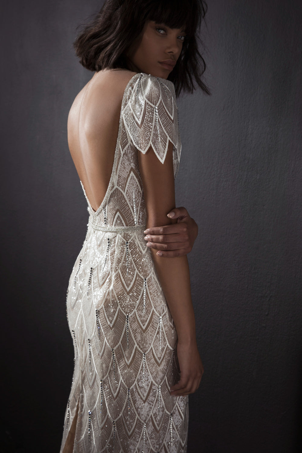 אישה עם תספורת קצרה וכהה לובשת את שמלת הכלה הנוצצת "Bridal Collection 2017 - Kimber", שגבה הפתוח ודוגמאות הארט דקו משקפים את האלגנטיות של שמלות כלה על רקע כהה ומרקם.