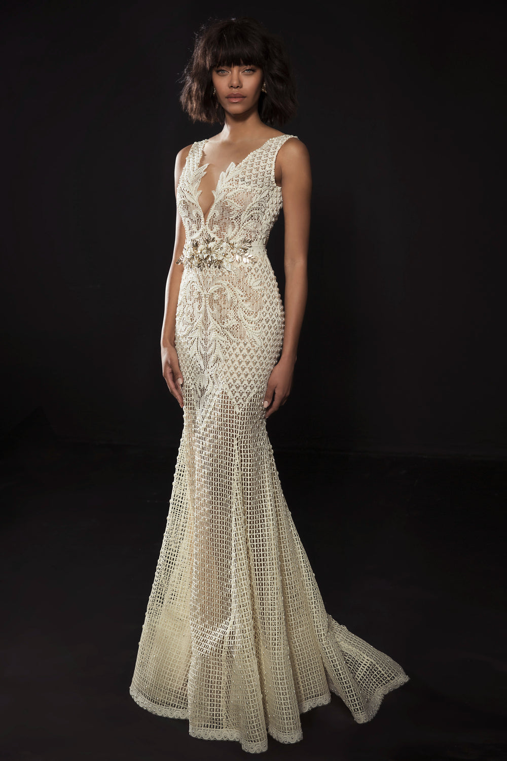 אישה עומדת על רקע כהה לבושה בשמלת קסידי מקולקציית הכלות 2017 של דרור קונטנטו, עם תחרה אלגנטית, עיטורי חרוזים, מחוך שקוף עם מחשוף עמוק וקישוטים פרחוניים במותניים.