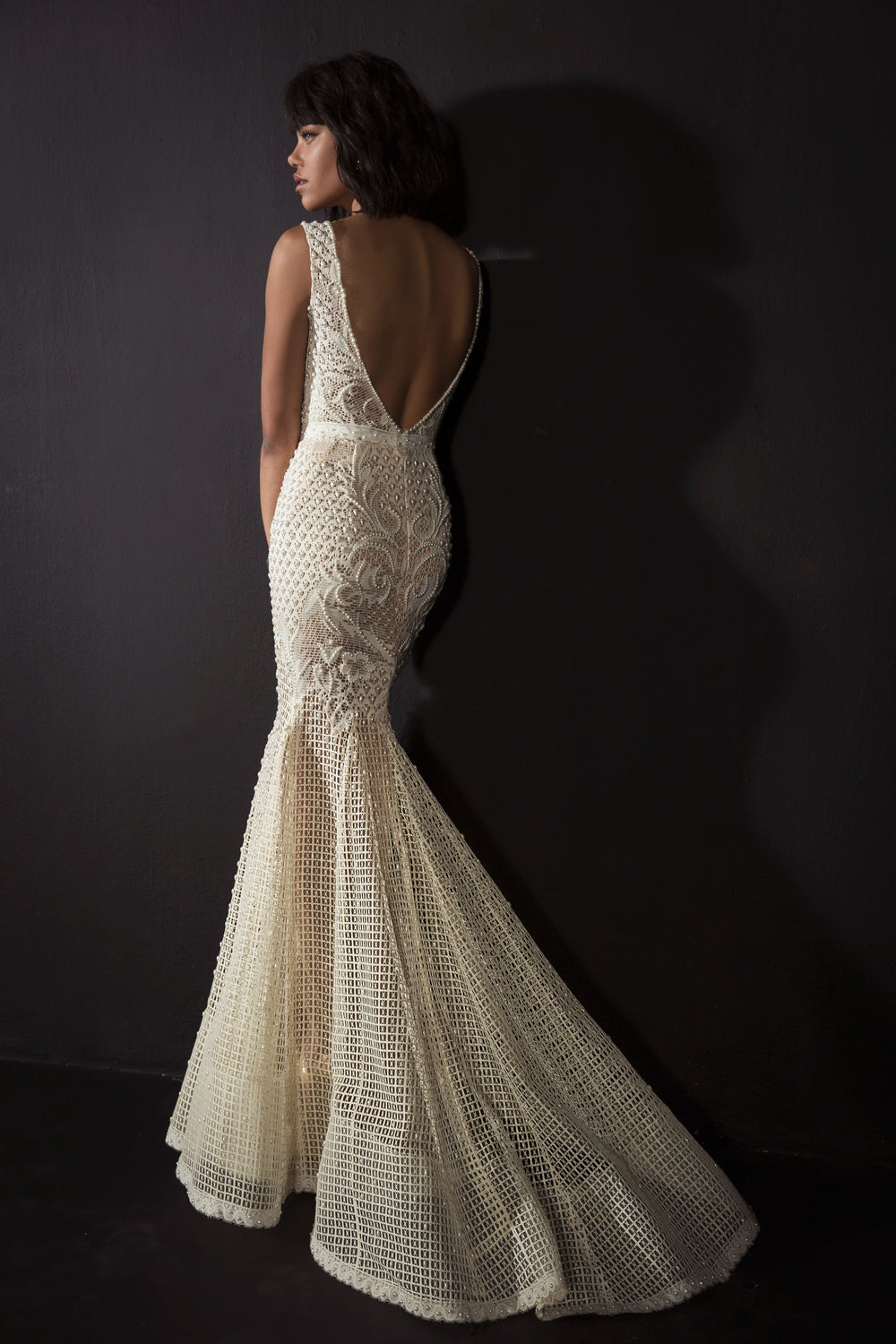 אישה עם שיער קצר וכהה עומדת על רקע כהה בשמלת קסידי מקולקציית הכלות 2017 של דרור קונטנטו — שמלת סטרפלס צמודה בצבע שנהב עם תחרה, בעלת דוגמאות מורכבות וגב עמוק בצורת V.