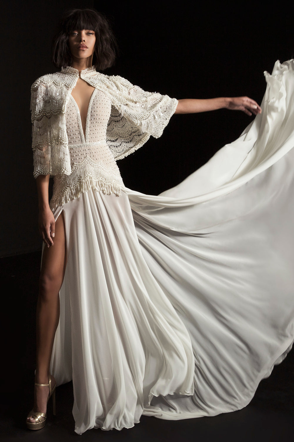 אישה בשמלת "לינה" מקולקציית הכלות 2017 של דרור קונטנטו, עם מחשוף עמוק, חתך עד הירך וגימור מעוטר, עומדת עם זרוע אחת מורמת, פורשת את הבד על רקע כהה.