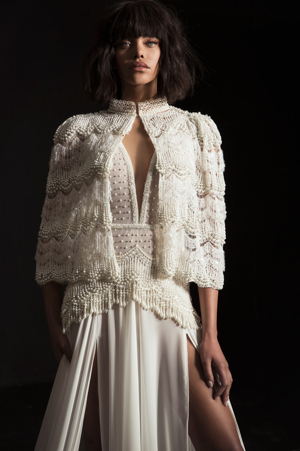 אדם עומד על רקע כהה ולובש את שמלת הכלה של דרור קונטנטו מקולקציית 2017 - לינה: שמלת כלה לבנה וזורמת עם חתך גבוה, עם גלימה משובצת חרוזים, עיטורים דמויי פנינים ופרטי רשת שקופים. שמלות כלה אלגנטיות מחודשת.