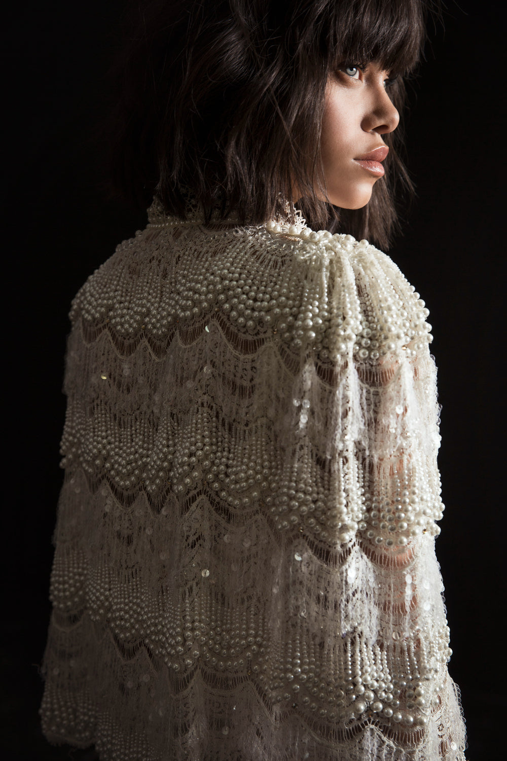 אישה עם שיער קצר וכהה מביטה לצד, לבושה בשמלת לינה בצבע שמנת, מעוטרת בחרוזים ותחרה, מקולקציית הכלות 2017, עם דוגמאות מסולסלות על רקע כהה.