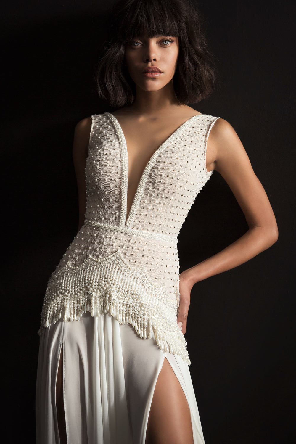 אישה עומדת על רקע כהה לבושה בשמלת "Bridal Collection 2017 - Lina", שמלת לבנה ללא שרוולים בעיצוב דרור קונטנטו, עם מחשוף V עמוק, עיטורי פנינים וחצאית מתנפנפת עם חתכים גבוהים.