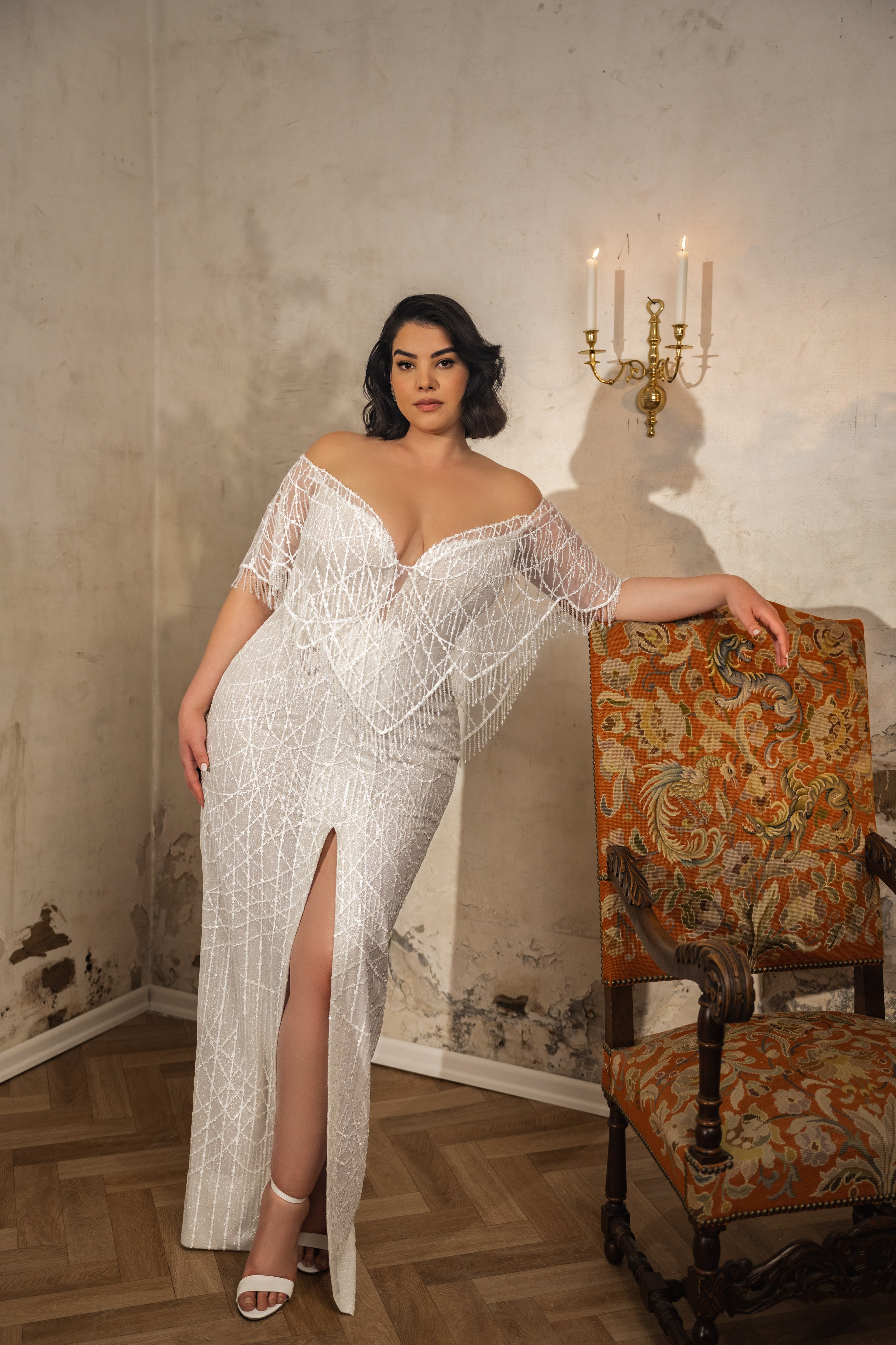 אישה מקולקציית Bridal Plus 2023/24 - שמלת Diana, עם תחרה לבנה נוצצת, עיטורי קריסטל, מחשוף כתפיים חשופות וחתך עד הירך, עומדת ליד כיסא מעוטר בחדר וינטג' עם קירות מרקמים ופמוט.