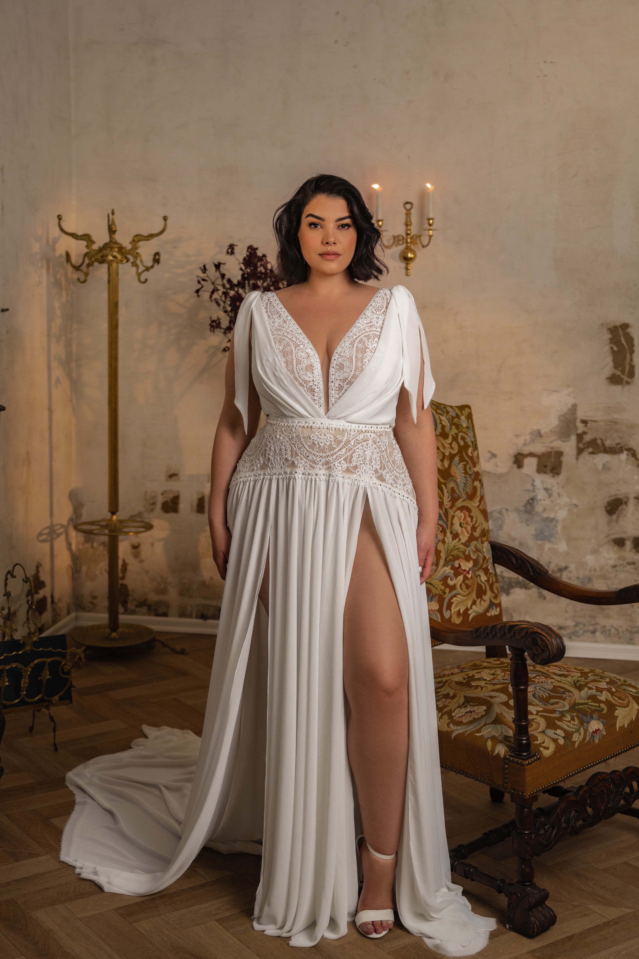 אישה לבושה בשמלת תחרה עם חתך גבוה מהקולקציה "Bridal Plus Collection 2023/24 - Kristen" עומדת בתוך הבית ליד כיסא עץ מעוטר, עם קיר מרקם, נברשת ומתלה מעילים ברקע.