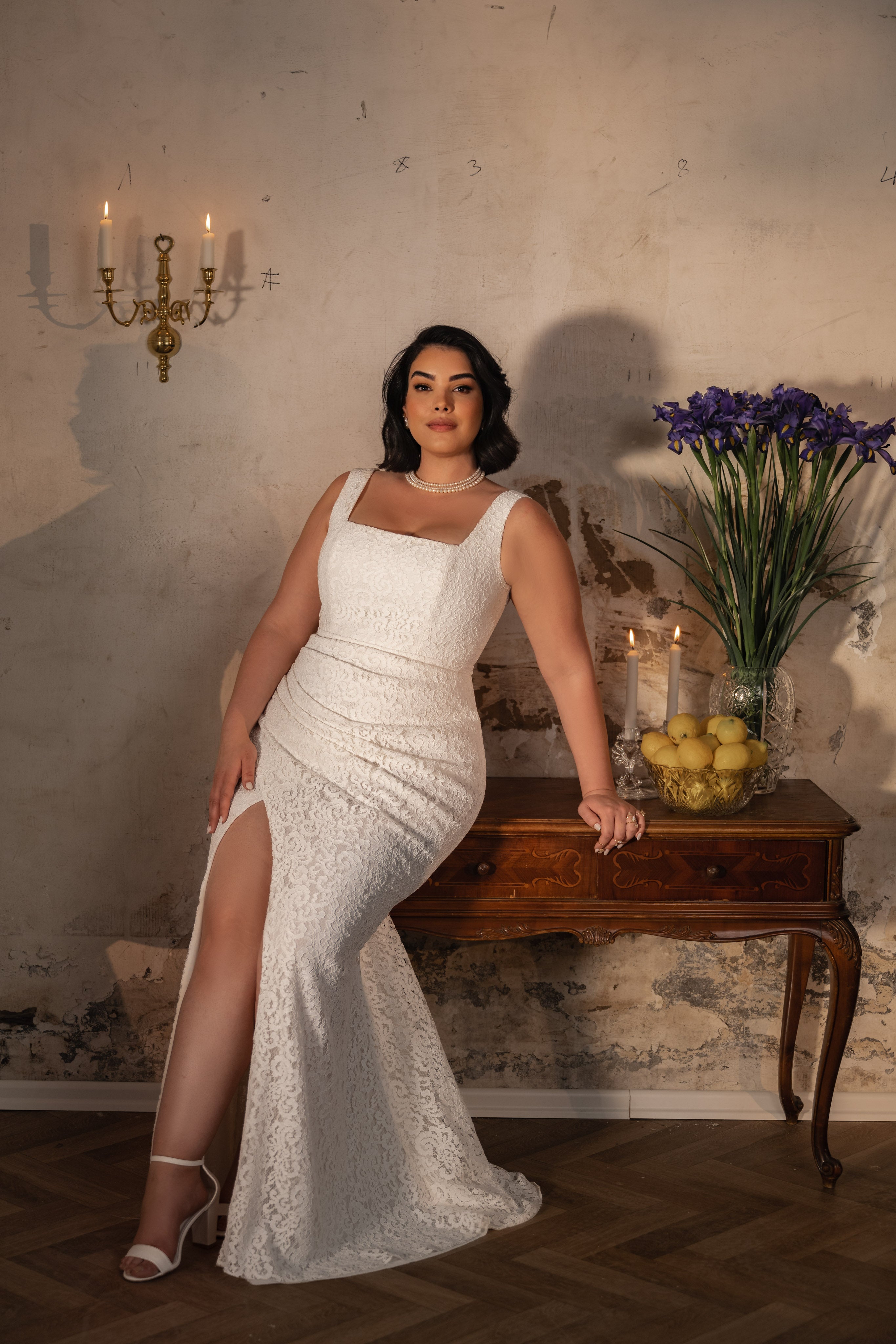 אישה מקולקציית Bridal Plus 2023/24 - שמלת Irene, עם תחרת פרחים לבנה וחתך גבוה עד הירך, מצטלמת ליד שולחן עץ עם נרות, לימונים ופרחים סגולים בחדר מואר באור רך עם עיצוב וינטג'.