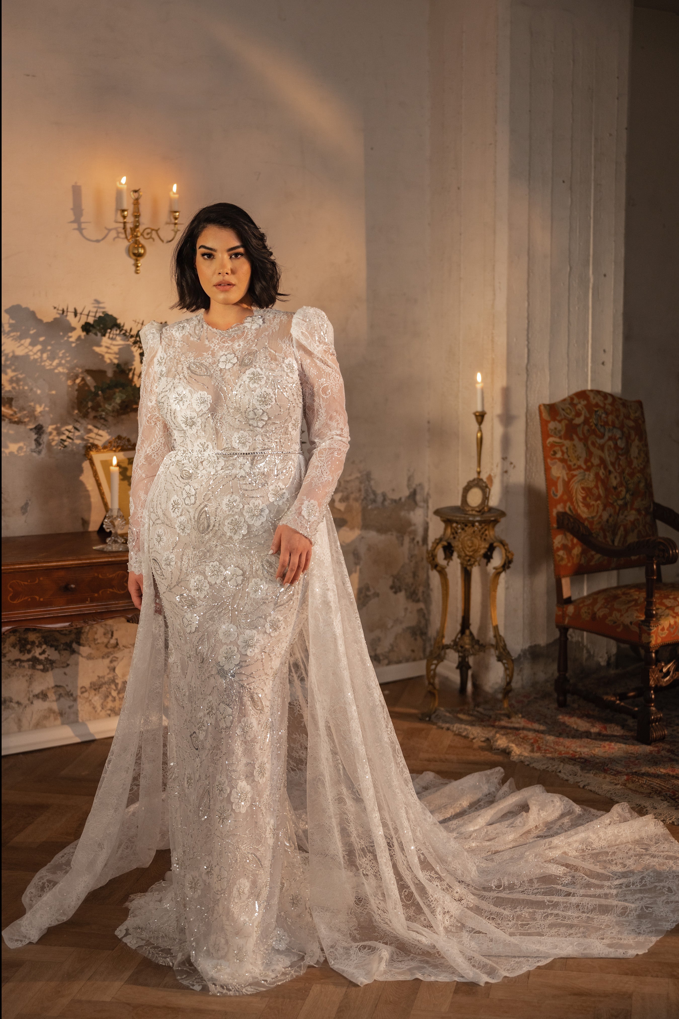 אישה לבושה בשמלת הכלה Shelby מקולקציית Bridal Plus 2023/24, שמלת תחרה ארוכה ומנצנצת עם שרוולים שקופים ושובל דרמטי, עומדת בחדר וינטג' אלגנטי המעוטר בנרות, רהיטים עתיקים וקישוטים פרחוניים.