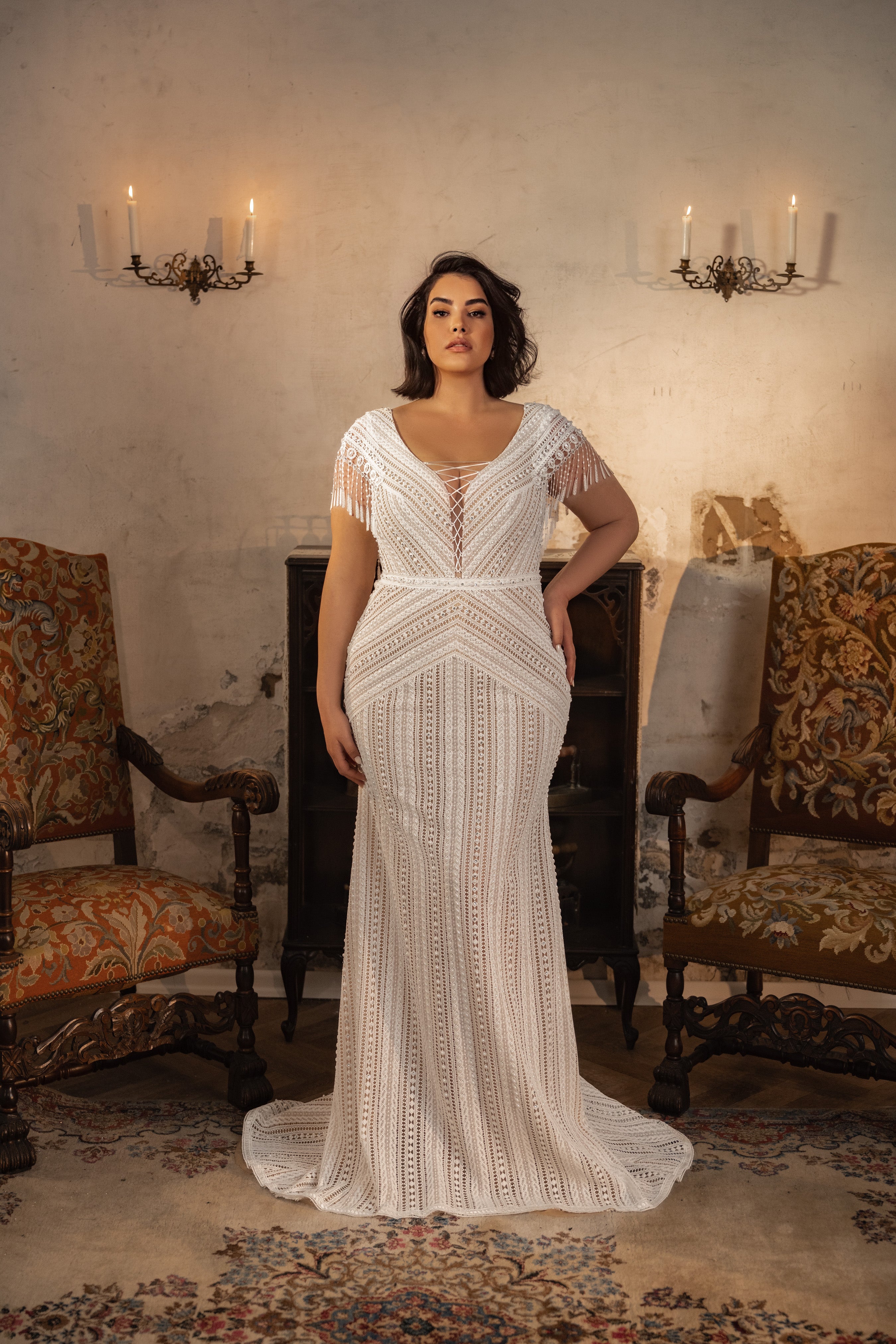 אישה מקולקציית Bridal Plus 2023/24 - מרטינה, שמלה לבנה עם תחרה באורך מלא, עומדת בין שני כיסאות מעוטרים בחדר וינטג' עם פמוטי נרות ושטיח עתיק.