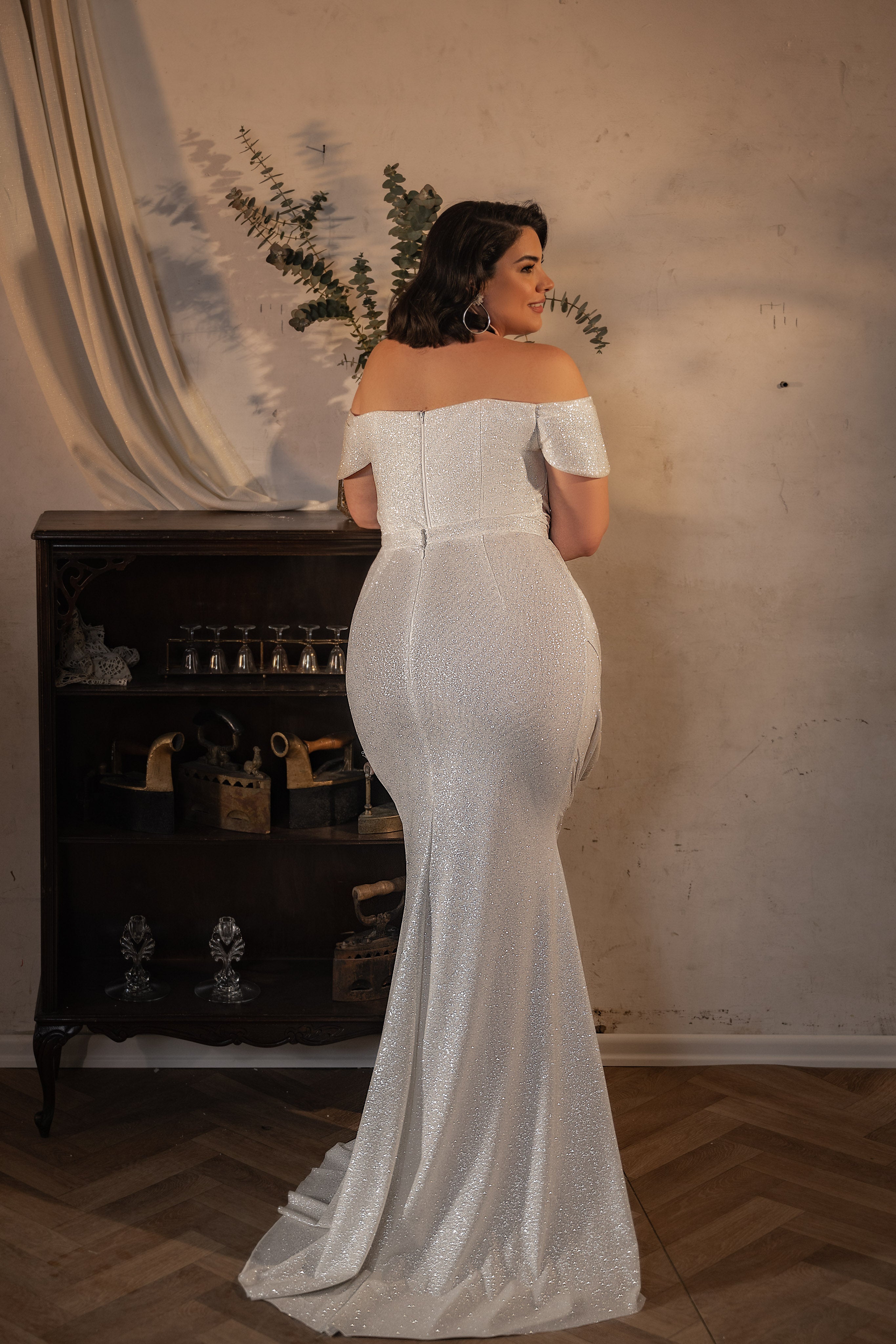 אישה מקולקציית Bridal Plus 2023/24 - שמלת כלה לבנה באורך מלא עם כתפיים חשופות מבית Taylor עומדת עם גבה למצלמה בחדר מואר באור חם, לצד ארון עץ כהה עם פריטי נוי ואגרטל עם צמחייה.