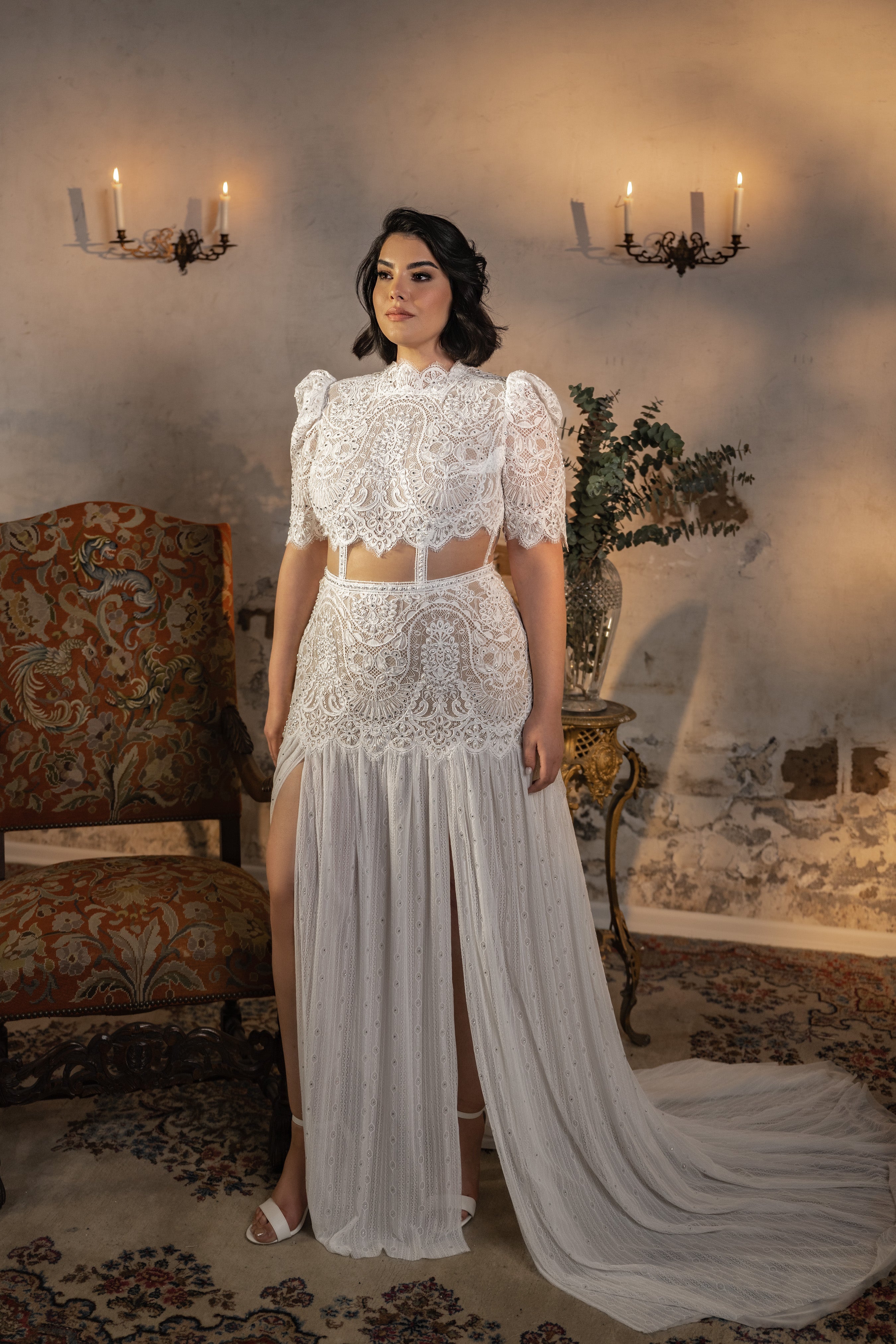 אישה מקולקציית Bridal Plus 2023/24 - Albi, שמלת כלה לבנה מתחרה במידות גדולות עם חיתוכים בצדדים, חצאית שיפון וחתך גבוה, עומדת בתוך הבית ליד כורסה מעוטרת ונרות דולקים.