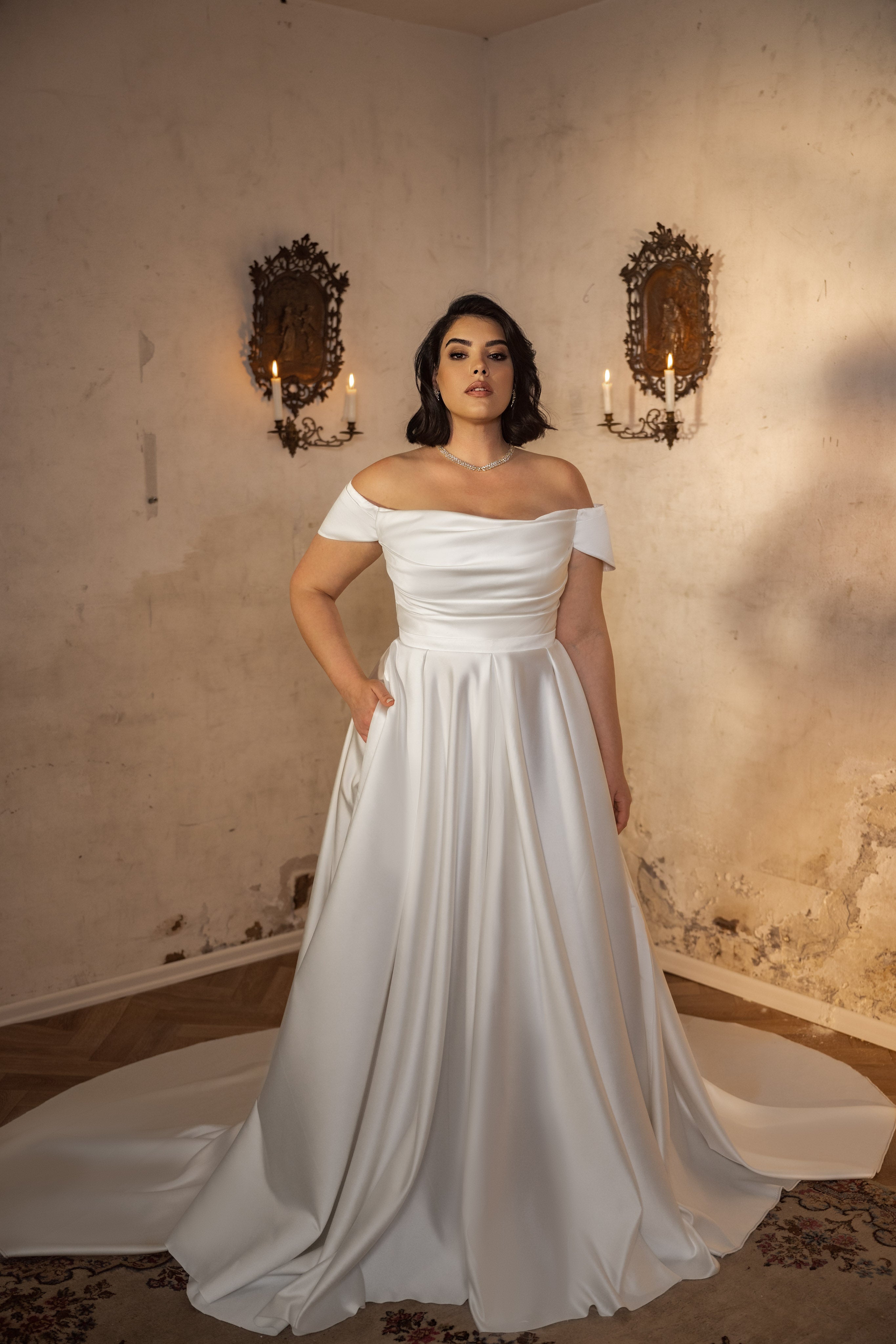 אישה לובשת את קולקציית Bridal Plus 2023/24 - Alicia, שמלת כלה לבנה עם כתפיים חשופות מבד סאטן, עומדת בחדר רומנטי עם קירות מרקמים מיושנים ושני פמוטים מעוטרים המוארים בנרות.