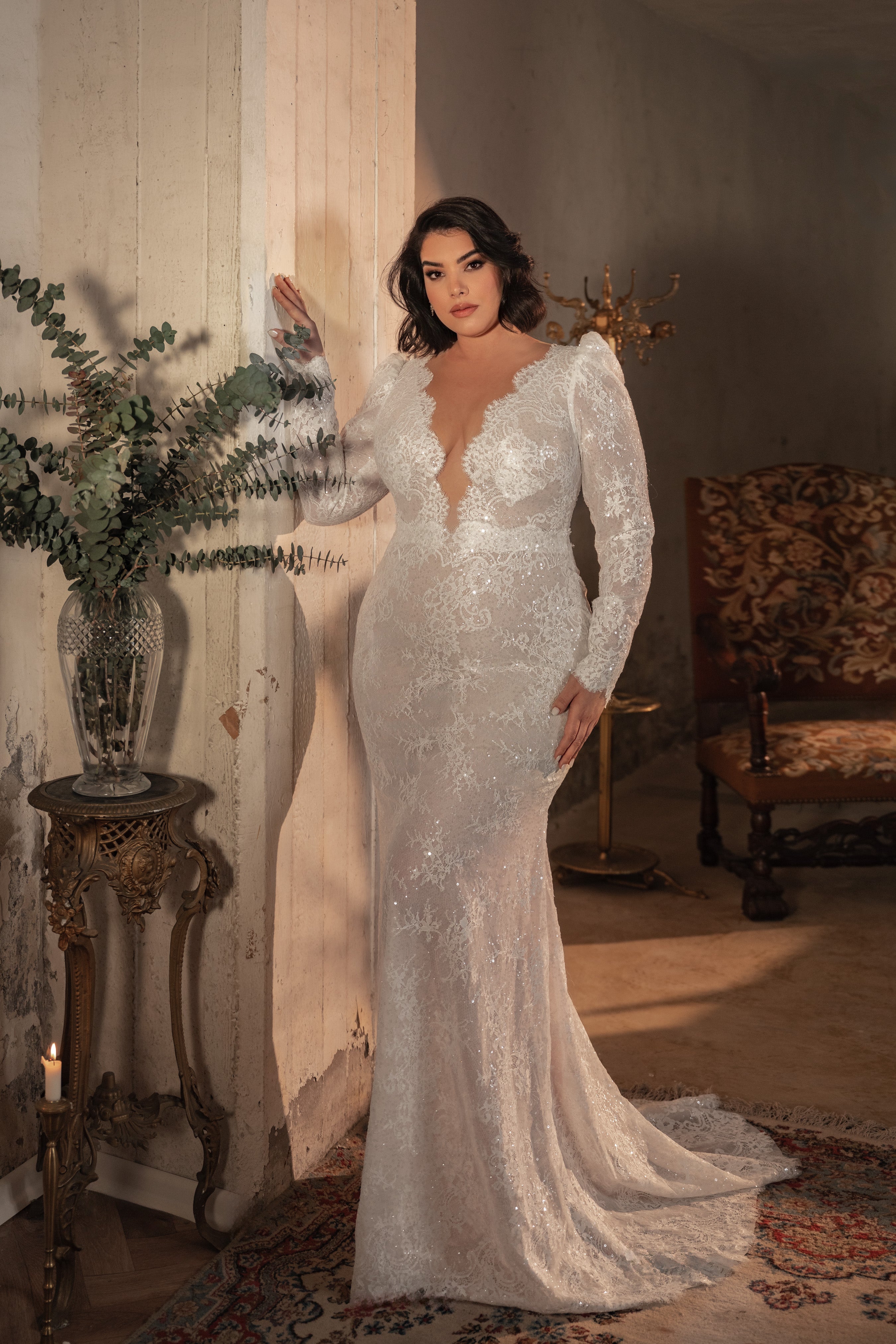 אישה מקולקציית Bridal Plus 2023/24 - שמלת כלה עם שרוולים ארוכים מתחרה Hope עומדת בתוך הבית ליד קיר עץ כפרי, עם שולחן וינטג', צמחייה וכיסא מעוטר ברקע.