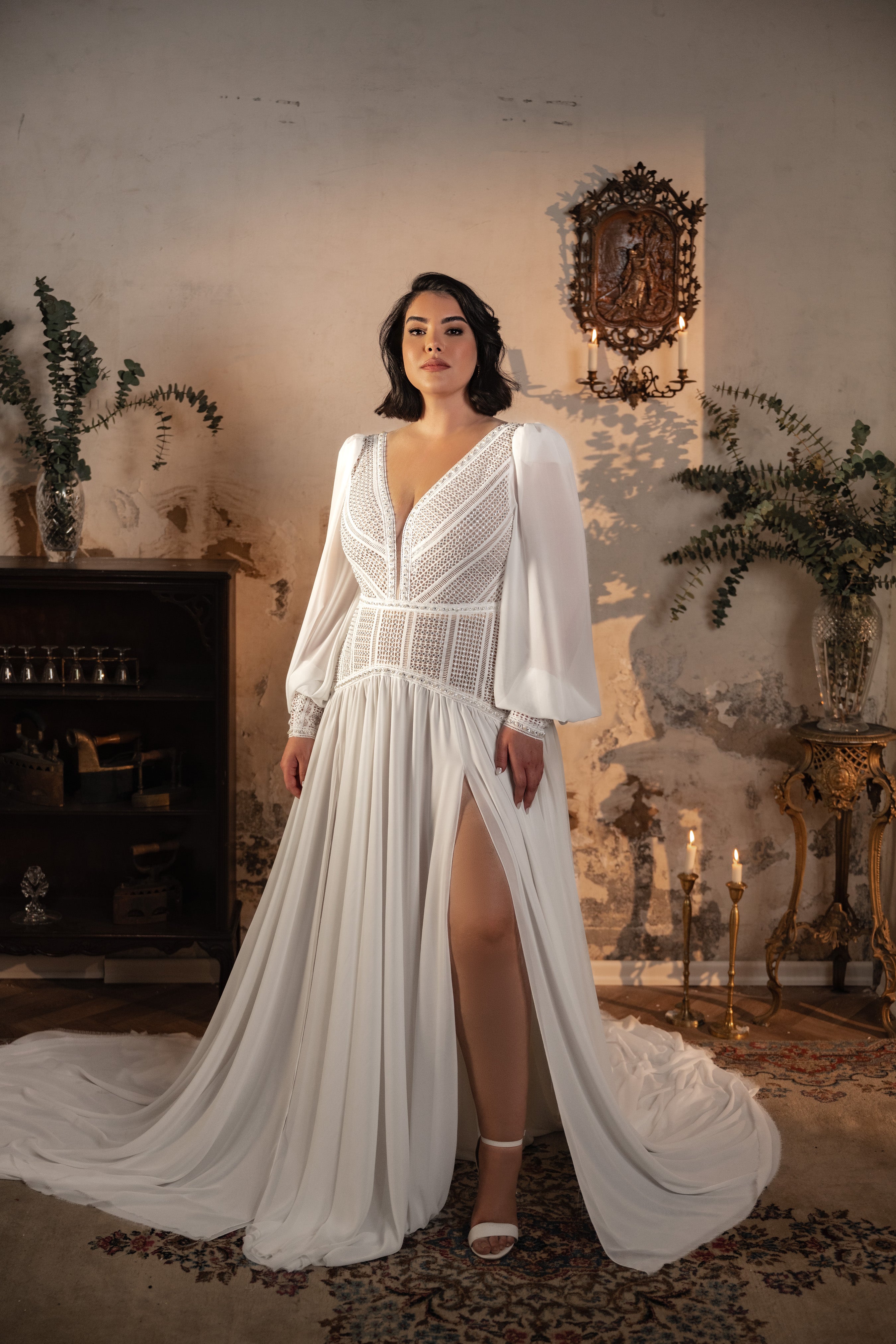 אישה מקולקציית Bridal Plus 2023/24 - שמלת Georgina, עם שרוולים ארוכים לבנים, מחשוף V עמוק וחתך גבוה, עומדת בתוך הבית על שטיח מעוצב עם נרות, צמחים ועיצוב כפרי על רקע קיר מרקם.