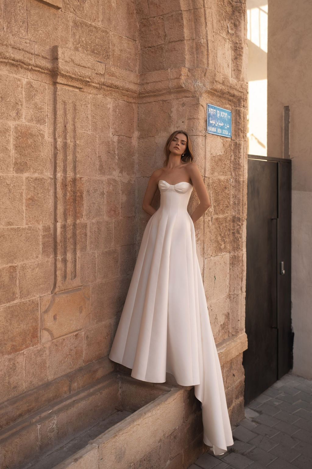 אישה לבושה במחוך סטרפלס, עוטה את שמלת הכלה NADYA W מקולקציית שמלות הכלה – MID 2026, עומדת על רקע קיר אבן מתחת לשלט רחוב עם כיתוב בעברית, בסביבה עירונית חיצונית מוארת בשמש — הצצה לאלגנטיות נצחית.