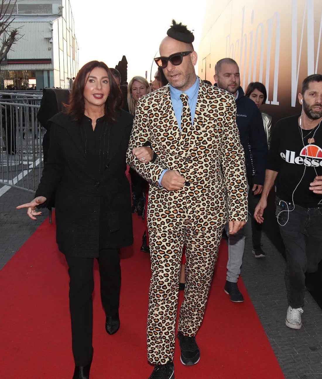 גבר בחליפה עם הדפס נמר וכובע קטן הולך בזרועות שלובות עם אישה לבושה בשחור על שטיח אדום, מוקפים באנשים אחרים. הם נמצאים בחוץ, עם בניינים וגדרות ברקע.