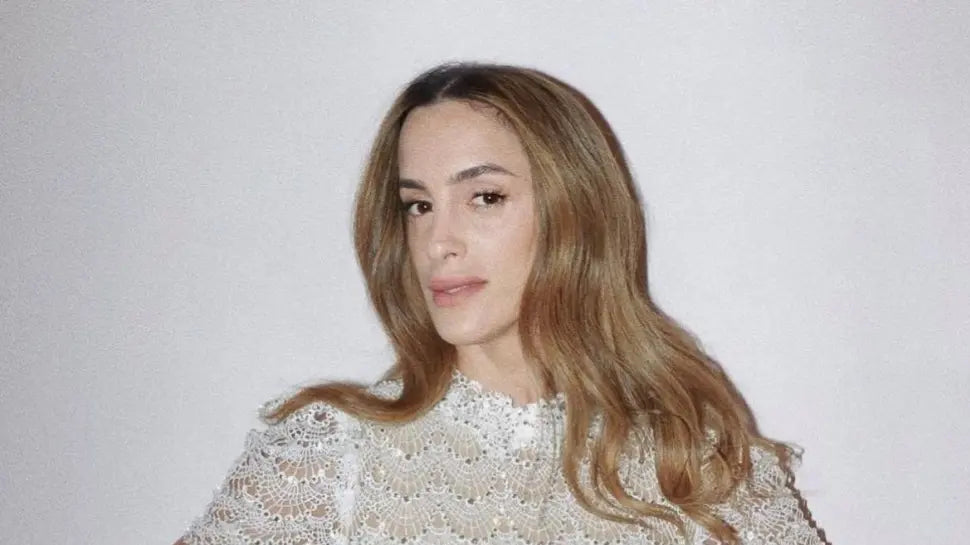 אישה עם שיער חום בהיר ארוך וגלי, לבושה בחולצת תחרה לבנה, עומדת מול רקע בהיר וחלקה, ומביטה בביטחון למצלמה.