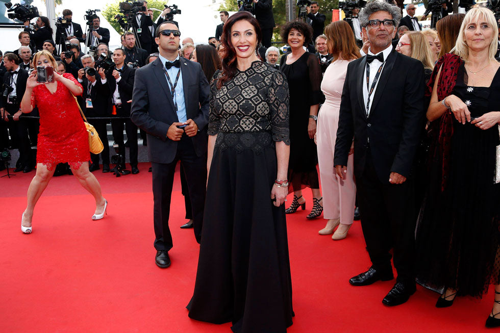 אישה בשמלה שחורה עומדת מחייכת על שטיח אדום, מוקפת באנשים לבושים בבגדים רשמיים וצלמים המתעדים את הרגע באירוע הומה אדם.