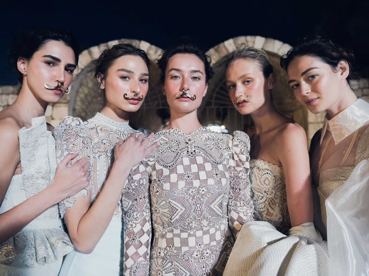 חמש נשים בשמלות מעוטרות וצבעוניות פוזות יחד בחוץ בלילה, כל אחת מהן חובשת שפם מזויף ומשעשע. ברקע נראים קשתות אבן מוארות באורות.