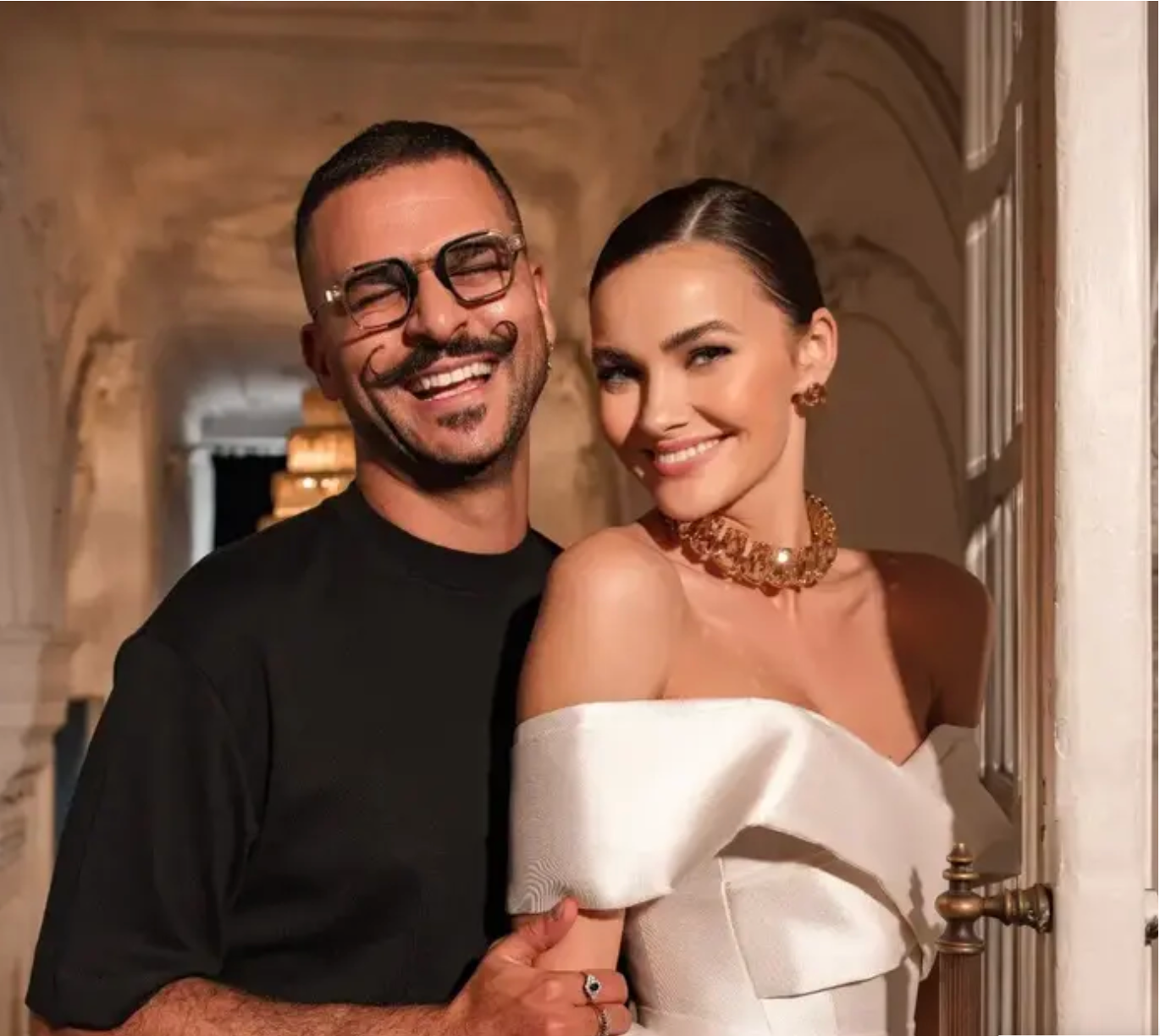 גבר עם משקפיים וחולצה שחורה עומד מחייך עם אישה בשמלה לבנה אלגנטית עם כתפיים חשופות וענק זהב. הם מצטלמים יחד בתוך חדר מואר באור חם, בסביבה מעוטרת.