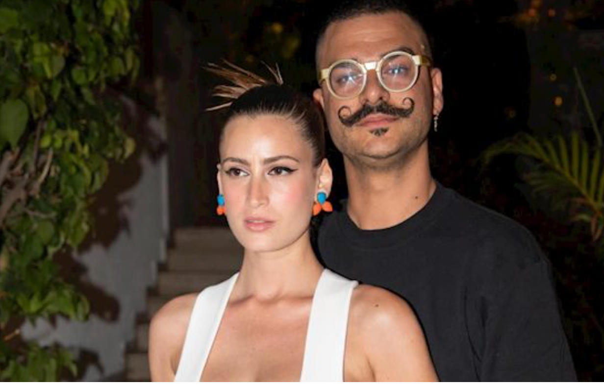 אישה עם שיער משוח לאחור ועגילים צבעוניים עומדת מול גבר עם משקפיים, עגילים ושפם מסולסל. ברקע נראים צמחייה ותאורה רכה.