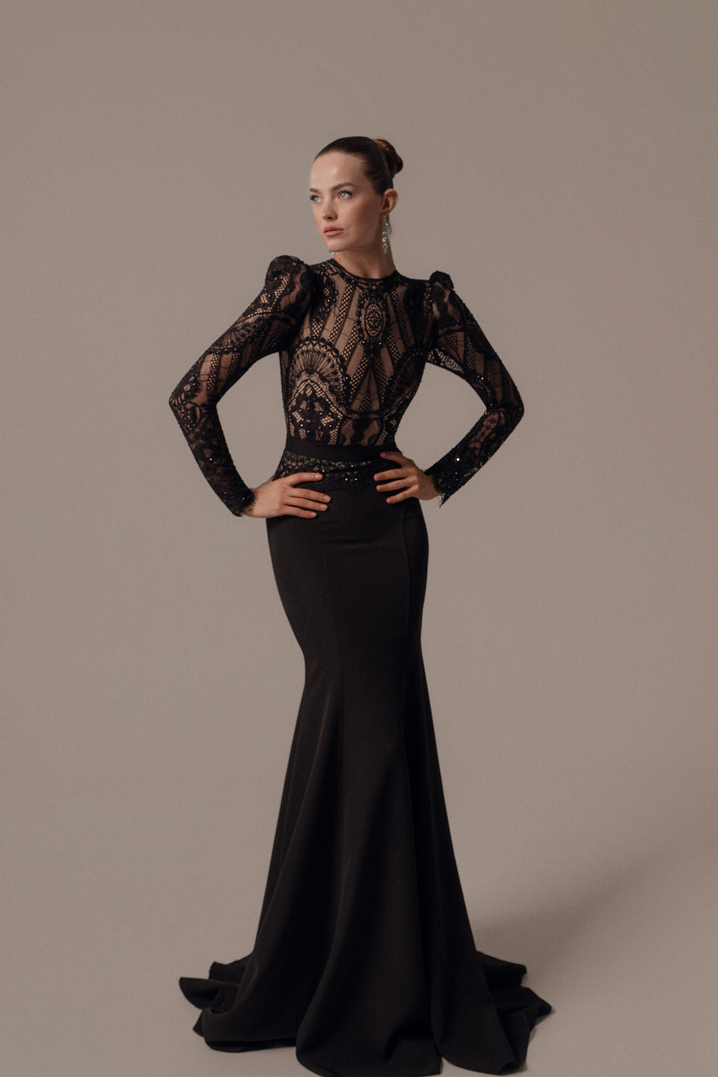 אישה משדרת ביטחון עצמי בשמלת Leya מקולקציית ערב 2026, שמלת ערב שחורה צמודה עם מחוך תחרה שקוף ומורכב ושרוולים ארוכים. היא מסדרת את שיערה לקוקו חלק עם עגילים מתנדנדים על רקע ניטרלי.
