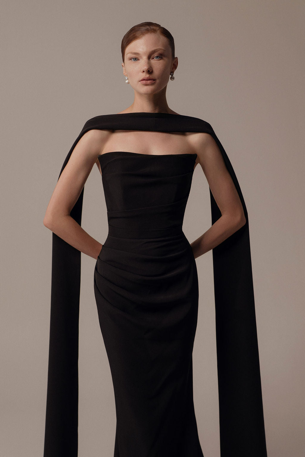 אישה עומדת בביטחון על רקע בז', לבושה בשמלת Nara מקולקציית הערב 2026 של דרור קונטנטו — שמלה שחורה אלגנטית ללא כתפיות עם כתף ארוכה ודרפית. היא משלבת אותה עם עגילי פנינה ושיער אסוף בקפידה.