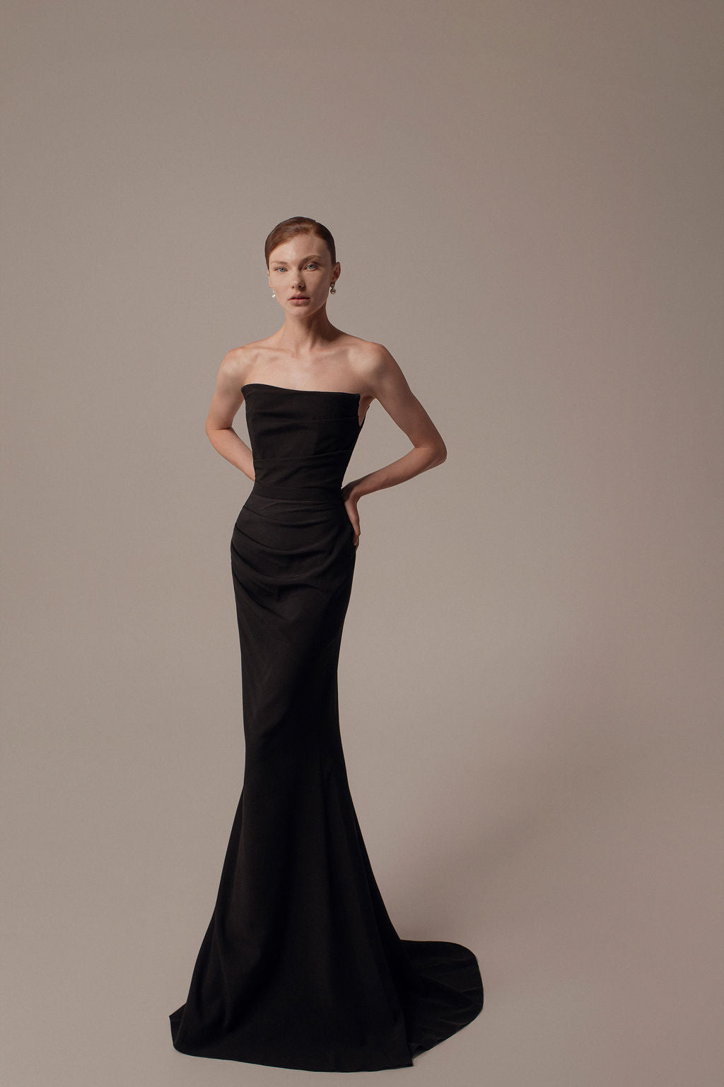 אישה עם שיער קצר מצטלמת בביטחון על רקע אחיד, לבושה בשמלת נרה שחורה צמודה ונטולת כתפיות מקולקציית ערב 2026 של דרור קונטנטו, באורך הרצפה עם שובל קל וידיה על מותניה.