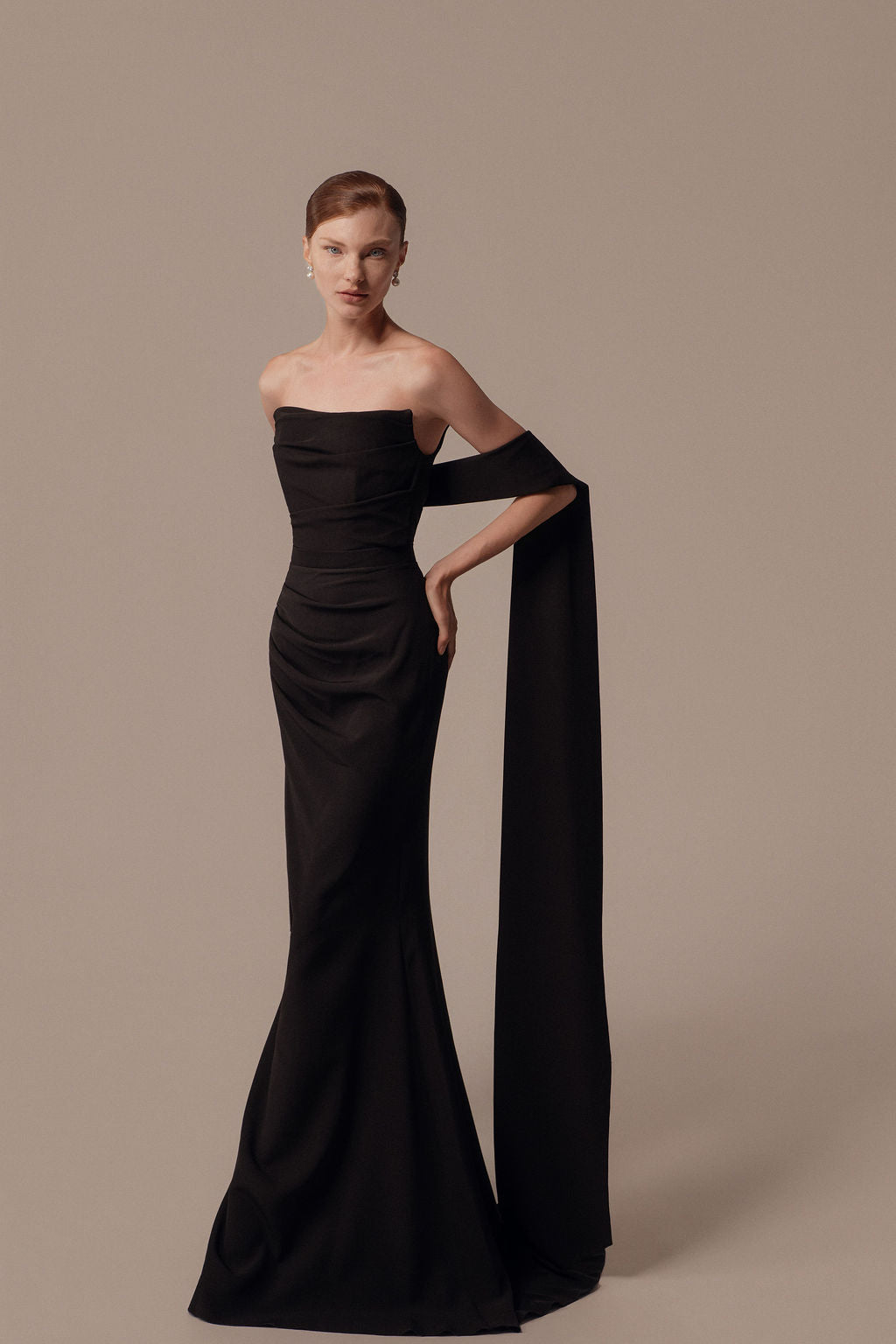 אישה עומדת על רקע אחיד לבושה בשמלת Nara מקולקציית הערב 2026 של דרור קונטנטו — שמלת ערב שחורה ללא כתפיות עם אבנט ארוך ודרפטי על זרוע אחת. שיערה מסודר והיא מצטלמת עם עגילי פנינה ויד אחת על מותנה.