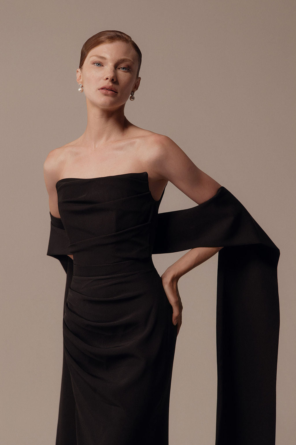 אישה בעלת עור בהיר ושיער קצר ומסורק לאחור מצטלמת עם יד אחת על המותן, לבושה בשמלת Nara מקולקציית Evening 2026 — שמלת ערב שחורה ואלגנטית עם אבנט ארוך ודרפטי, על רקע ניטרלי וחלקה.